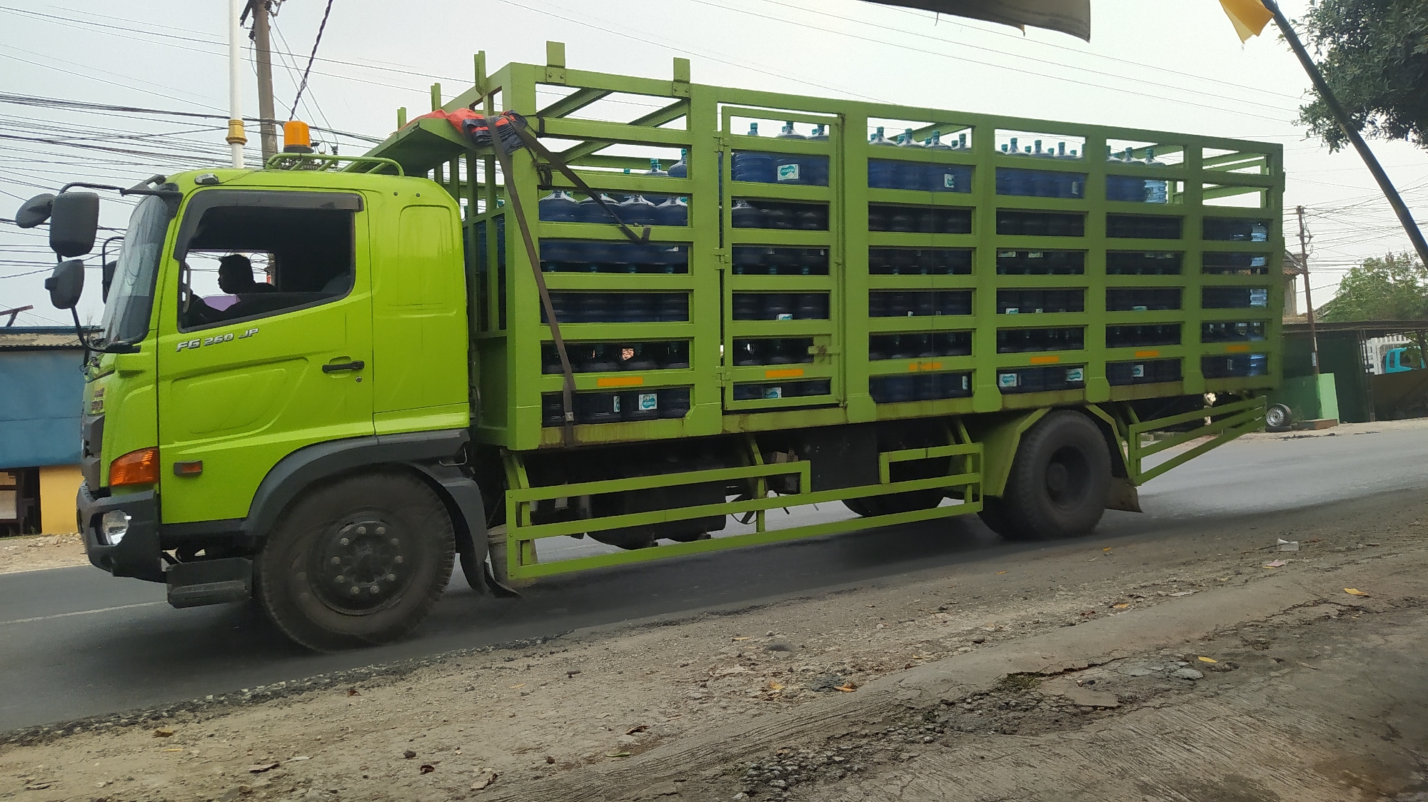 A bright lime green Hino FG 250 JP truck is parked on the side of a road in Cileungsi, Indonesia. The truck's cargo area is a tall green metal cage filled with rows of blue plastic water containers. The containers are stacked on multiple shelves within the cage, with at least four rows visible on each side. An orange safety light is mounted on the roof of the truck's cabin. Inside the cabin, the silhouette of a person, presumably the driver, is visible. The truck is positioned on a dusty, unpaved shoulder next to a paved road. The environment appears to be a semi-urban or industrial area, with some simple buildings and utility poles visible in the background. The sky is overcast, and the overall lighting suggests it is daytime.