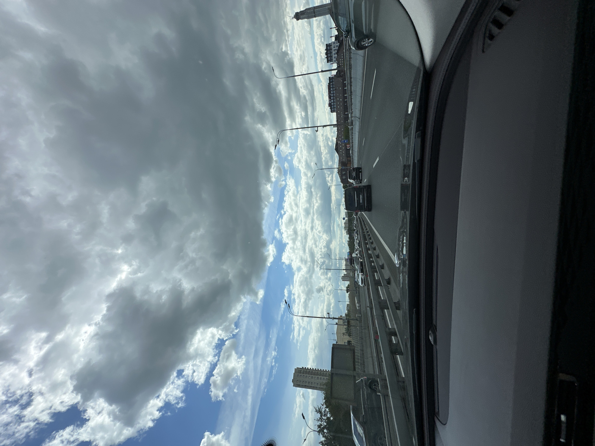The image shows a view from inside a car driving on a bridge in Moscow, Russia. The road is multi-lane and there are several other cars. The sky is partly cloudy. There are buildings visible on either side of the road. There are also streetlights along the bridge.