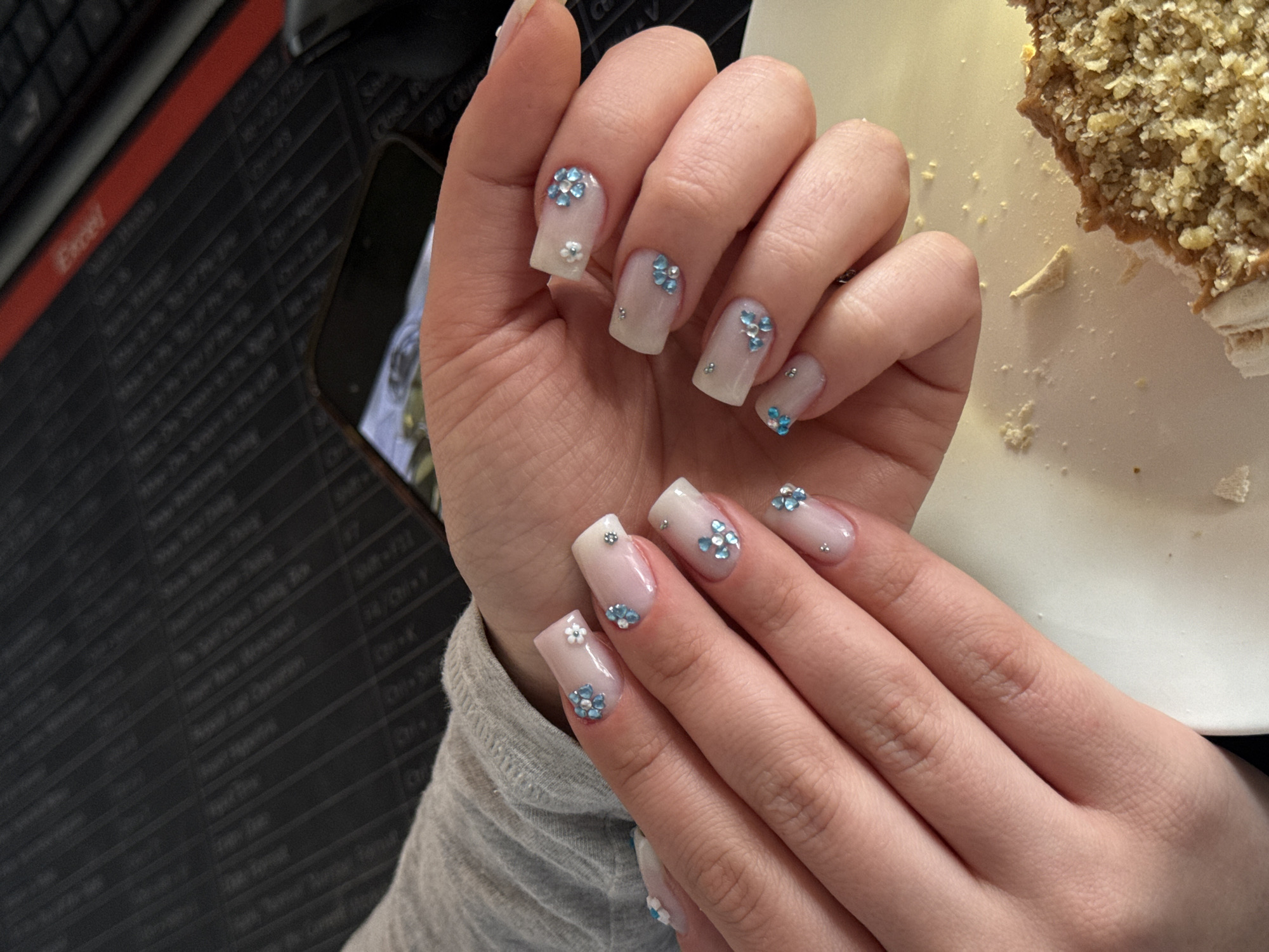 A close-up shot shows two hands with manicured square nails. The nails have a light pink base with small blue and white flower decorations. A keyboard and a smartphone lie on a black surface with white lettering. A partially eaten cake rests on a white plate beside the hands. The location is Yerevan, Armenia.