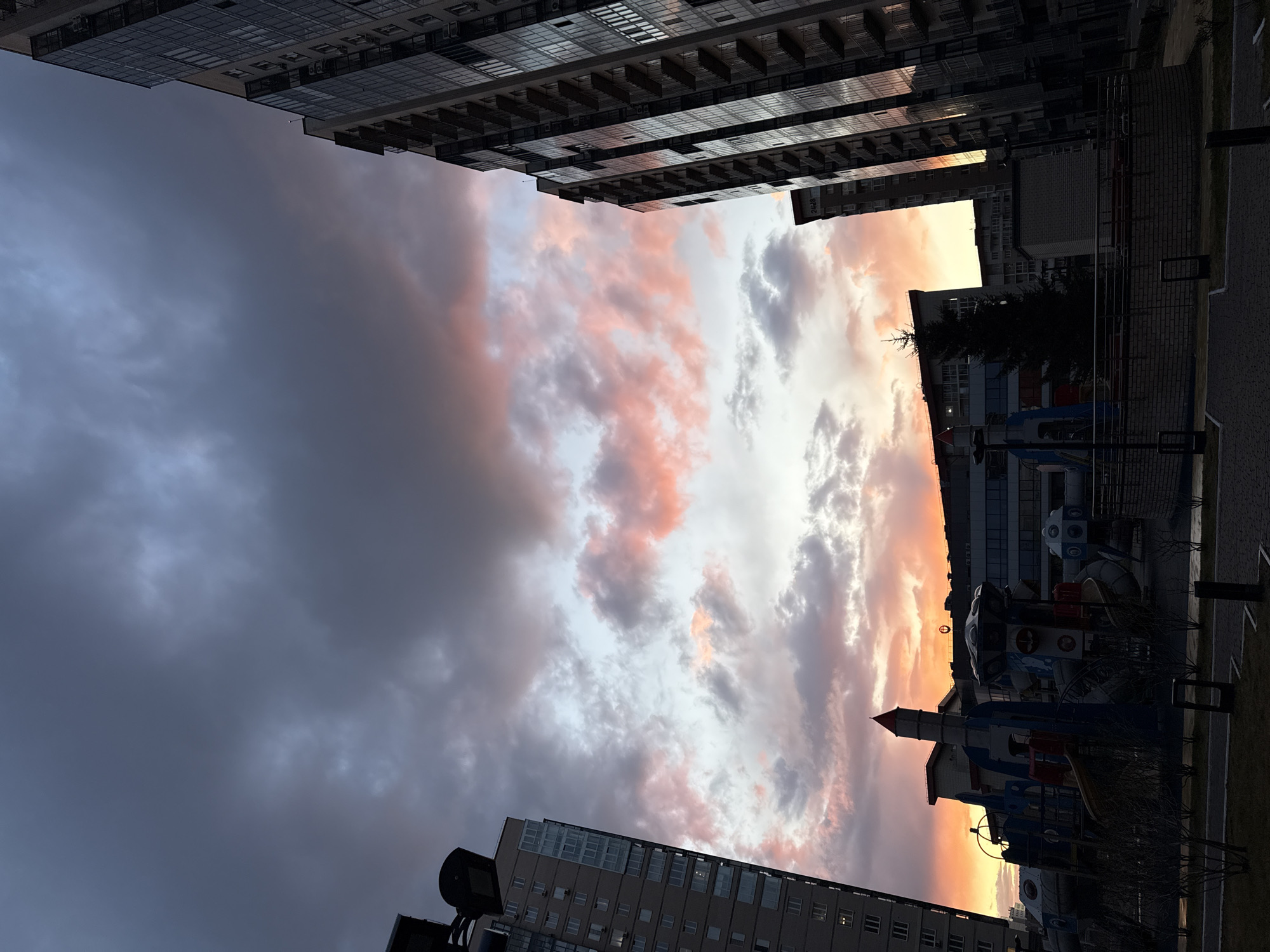 The image shows a view of the sky and surrounding buildings in what appears to be an urban setting. The sky is filled with clouds tinged with pink and orange hues, suggesting either sunrise or sunset. Tall buildings with multiple floors and reflective windows flank the sky on either side, creating a narrow, framed view.
In the lower portion of the image, there is a section of a playground with colorful structures, partially obscured by foliage. A tree and a fence can also be seen. The location is Krasnoyarsk, Russia.