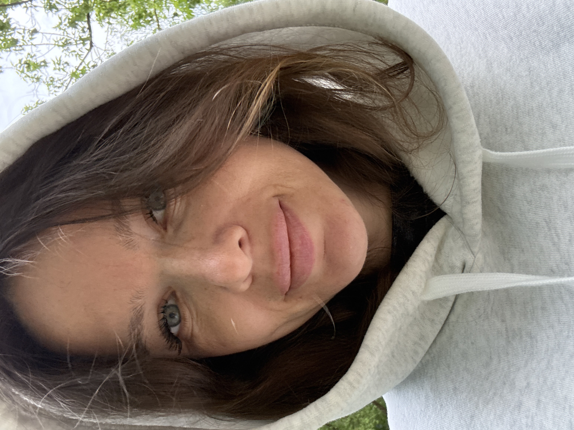 A woman with brown hair and light eyes wears a gray hooded sweatshirt. The hood is up, framing her face. She is looking at the camera with a slight smile. The background is slightly blurred, showing green foliage, suggesting she is outdoors. The location is not discernible as Moscow, Russia without additional visual or contextual information.