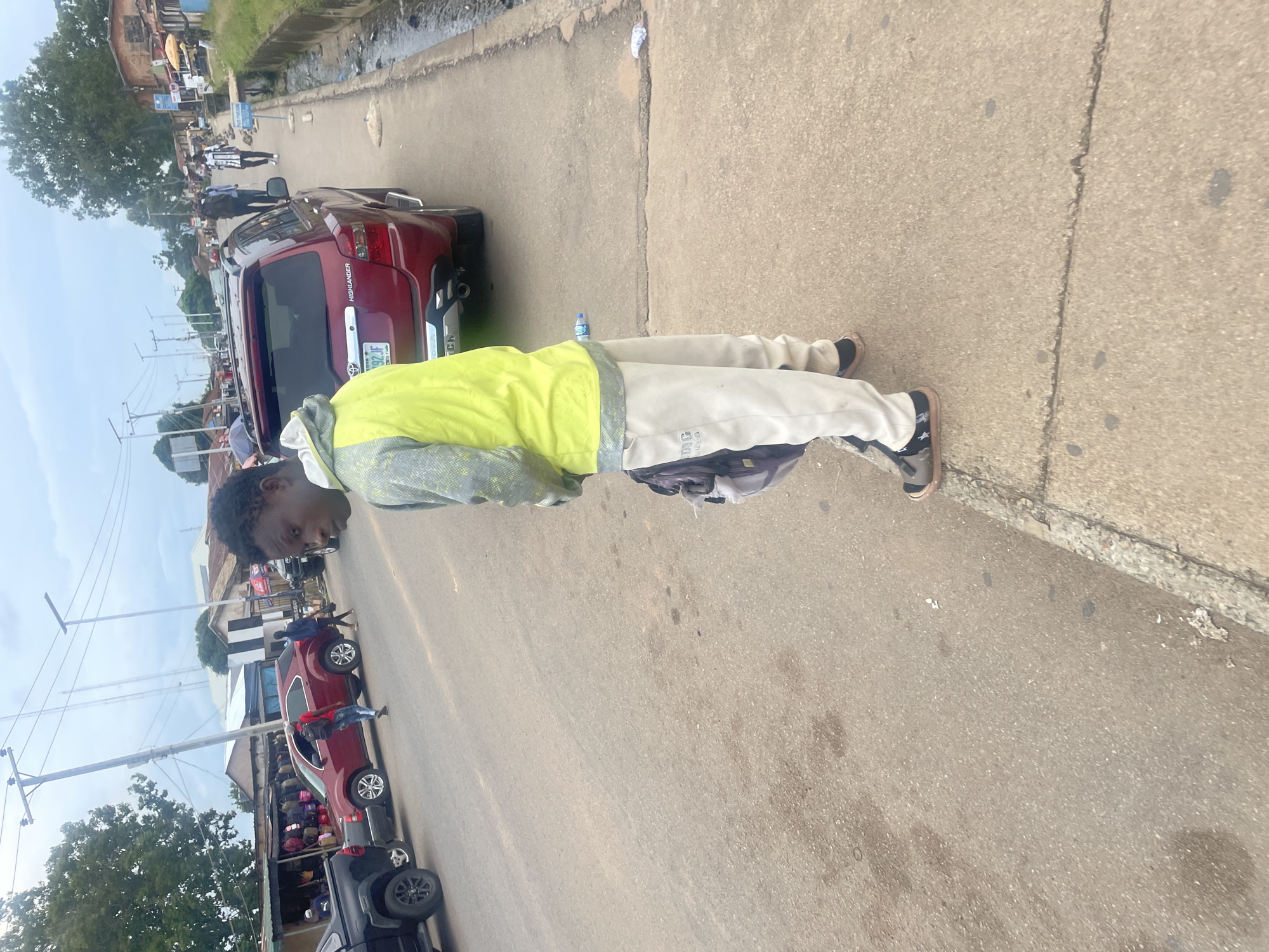 A young male, with dark skin and short dark hair, stands on a sidewalk next to a paved road. He is wearing a light-colored hooded jacket with gray accents and light-colored sweatpants. A dark backpack is slung over his left shoulder. His left foot is on the sidewalk, and his right foot is also on the sidewalk. He is looking towards the right and slightly back at the camera.
In the background, a red SUV is parked on the road, and a black pickup truck is visible further down the road to the left. There are several buildings and stalls lining the right side of the road, suggesting a market or commercial area. A narrow, debris-filled channel runs alongside the sidewalk on the right. The sky is overcast. The scene appears to be in an urban environment. Based on visual cues consistent with street scenes in Jos, Nigeria, the location is likely Jos, Nigeria.