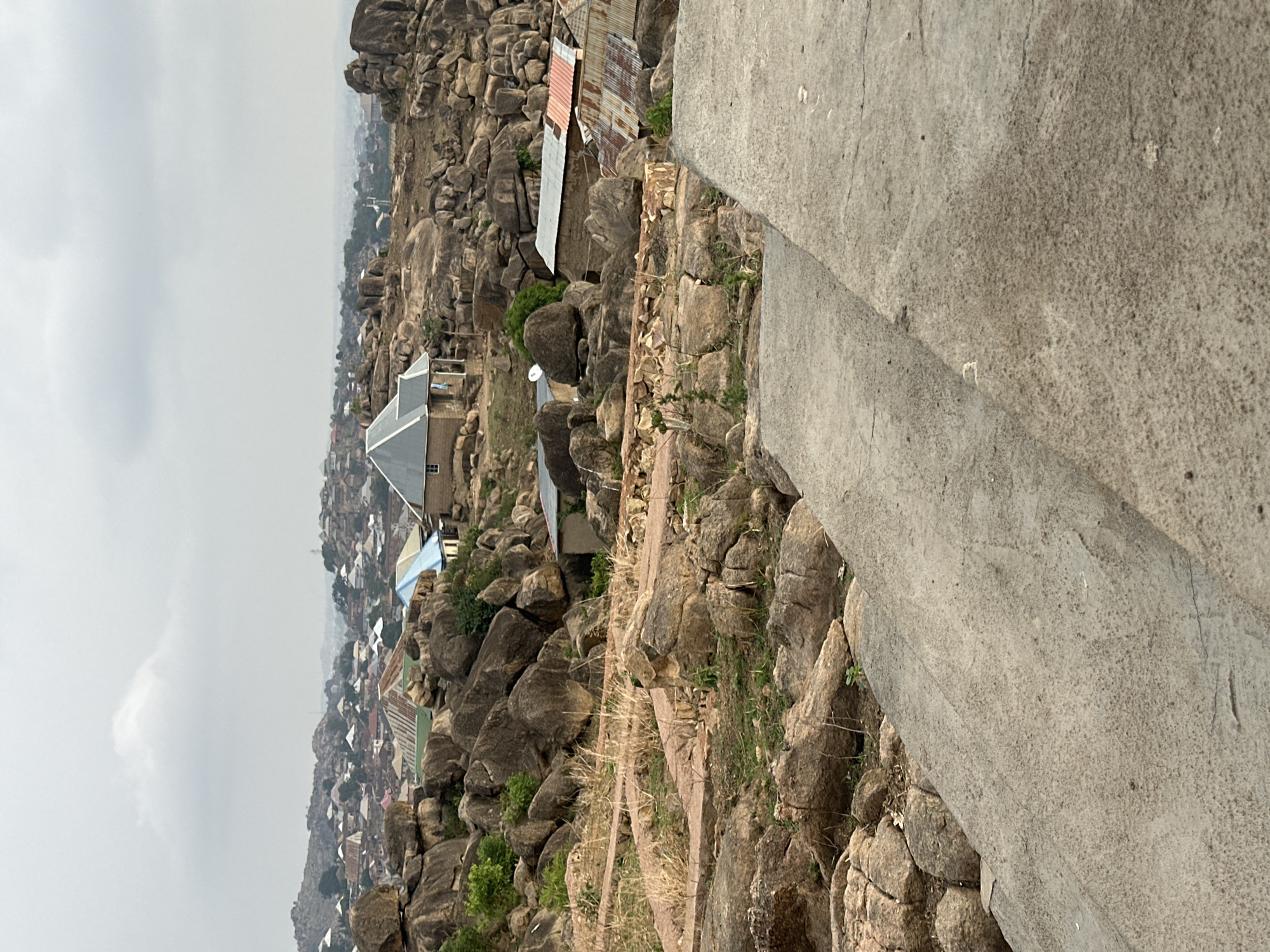 The image captures a section of the city of Jos, Nigeria, perched on rocky, elevated terrain. The foreground features a paved area with visible texture and cracks, suggesting a walkway or road. Beyond this, a cluster of natural rock formations dominates the landscape, interspersed with sparse vegetation. Several buildings with corrugated metal roofs are nestled among the rocks, indicating residential areas built into the natural landscape. In the distance, a dense urban settlement stretches towards the horizon, partially obscured by a hazy sky. The overall scene portrays a settlement uniquely integrated with a rocky, mountainous environment.