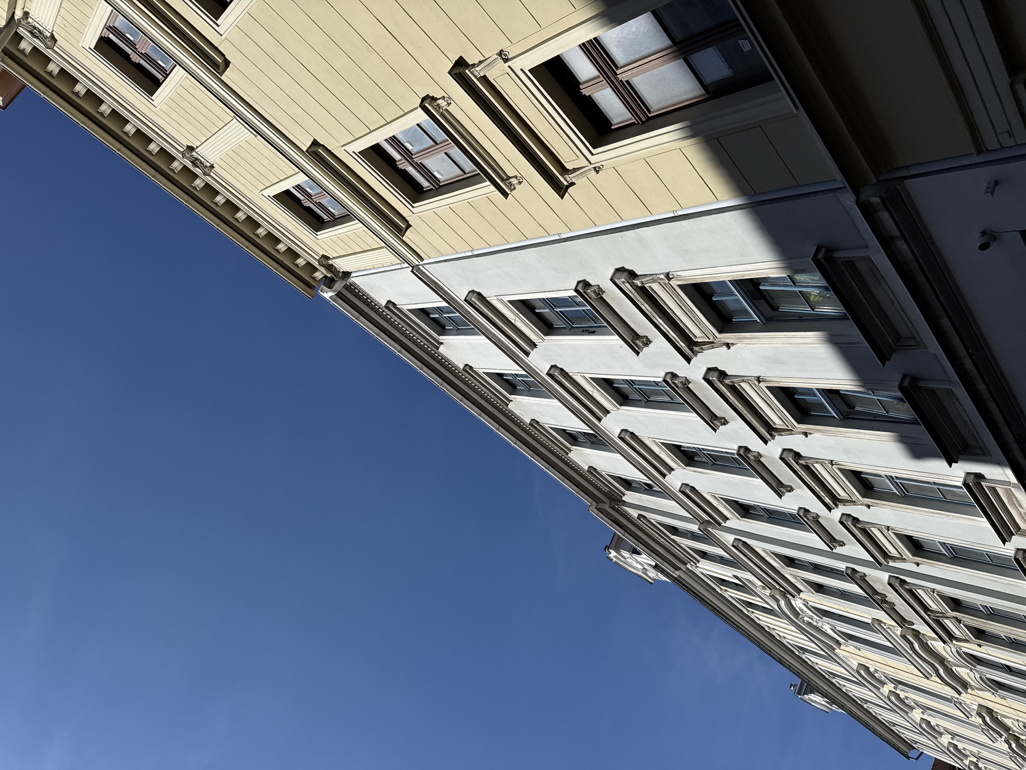This is a low-angle, full-frame shot of the facades of two adjacent buildings against a clear blue sky. The buildings are old European-style structures, with cream and pale yellow paint. They feature multiple stories with numerous windows. The building on the left is painted a light cream and has a row of windows with dark frames. The building on the right is painted a pale yellow and also has a series of windows with dark frames. Both buildings exhibit architectural details such as decorative moldings around the windows and cornices. Shadows are cast across the facades, indicating the direction of sunlight. The image appears to be taken on a sunny day. The specific location of these buildings in Görlitz, Germany, is not provided by the image alone.