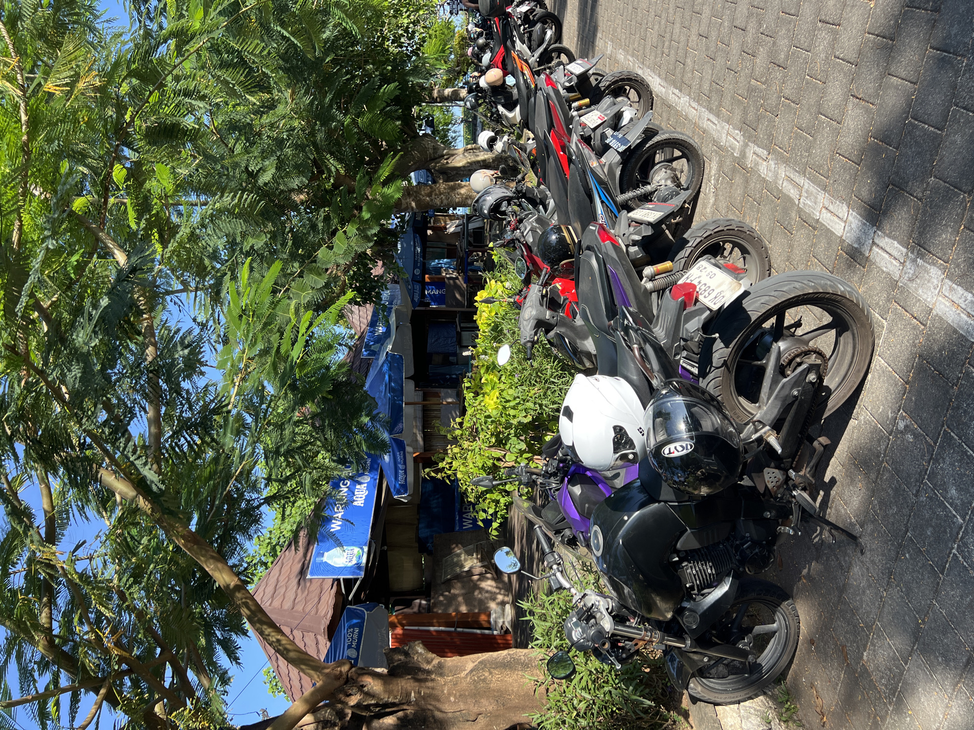 This photograph shows a row of motorcycles parked along a paved street in an urban setting. The motorcycles, varying in color from black and red to purple, are lined up parallel to the curb, each with a helmet placed on the seat or handlebars. The pavement is marked with a white line. A tree provides shade over the scene, its branches extending into the frame. A building, identified by signs advertising "WARLING AQUA" and "100% MURNI AQUA," suggests a local business along the street. The area appears to be a designated motorcycle parking zone in the city of Unknown, Unknown.