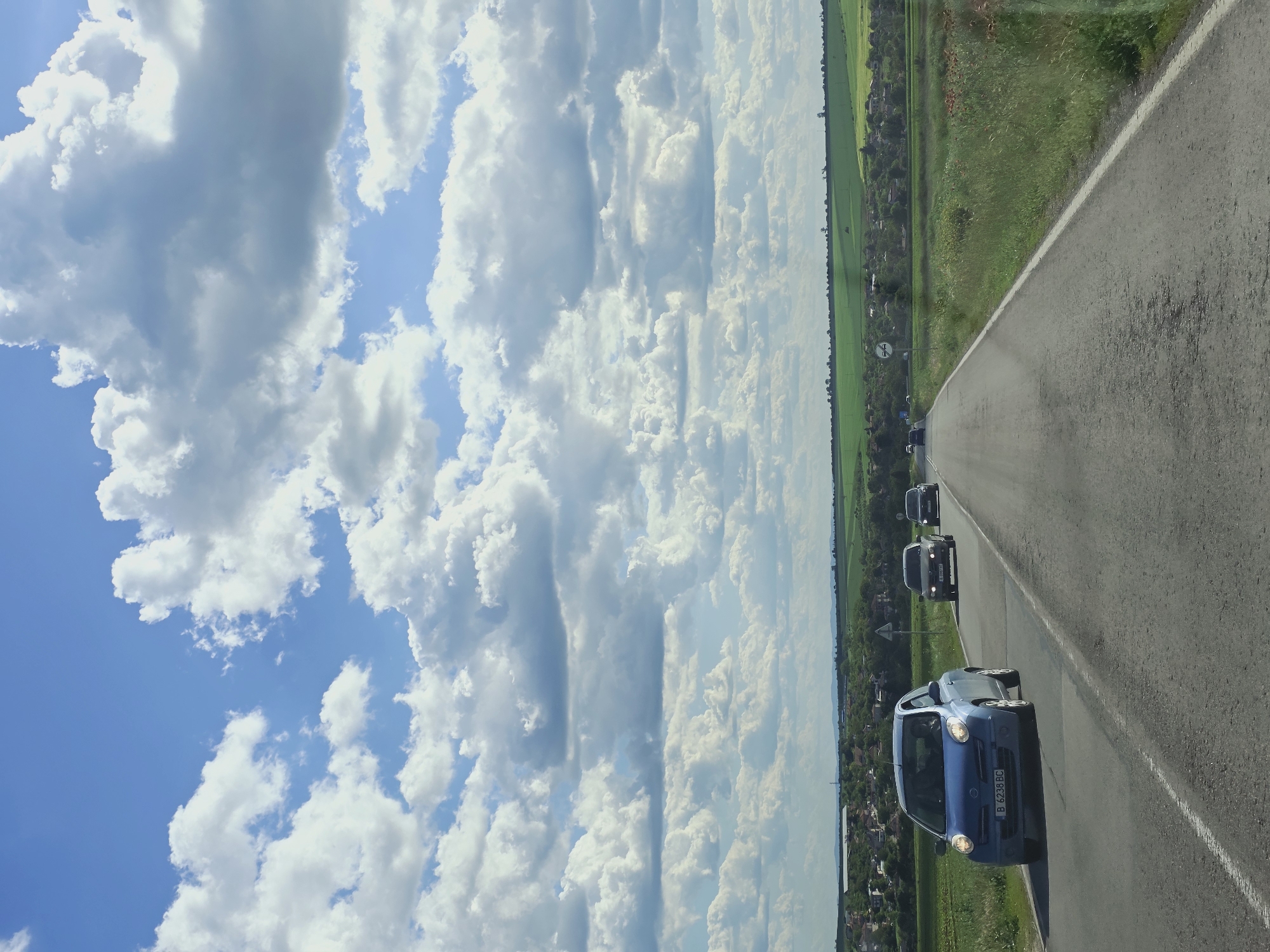The image depicts a rural road in Bulgaria, seemingly near the city of Kumanovo, as the license plate "B 6238 BC" is visible on the lead blue car, indicating registration in that area. Several vehicles are moving along the road, with the blue car in the foreground. The road is bordered by green fields on one side and a line of trees and a village on the other. The sky is predominantly cloudy, with patches of blue visible.