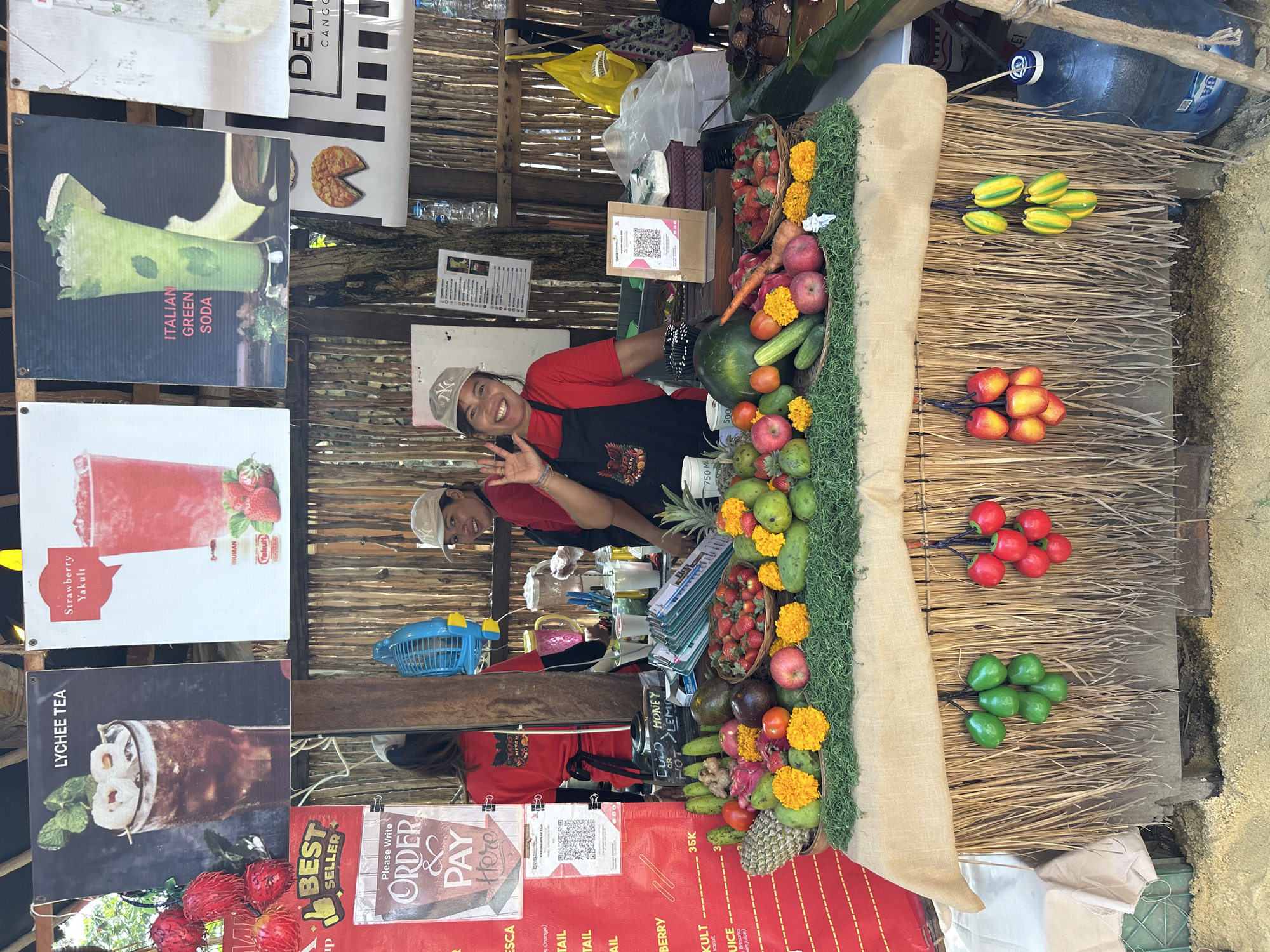 In North Kuta, Indonesia, a fruit stand displays a vibrant array of fresh produce, including strawberries, mangoes, dragon fruit, apples, avocados, cucumbers, and carrots, artfully arranged on a burlap-covered counter. Two women, both wearing red tops and aprons, stand behind the counter. One woman, in the foreground, waves at the camera with a broad smile, while the other, to her left, is partially obscured. Above the stand, colorful posters advertise various drinks, such as "Lychee Tea," "Strawberry Yakult," and "Italian Green Soda." The stand itself is constructed from natural materials like bamboo and thatch, giving it a rustic, tropical ambiance. In the background, large water dispensers and bags are visible, suggesting the operation of a bustling marketplace.