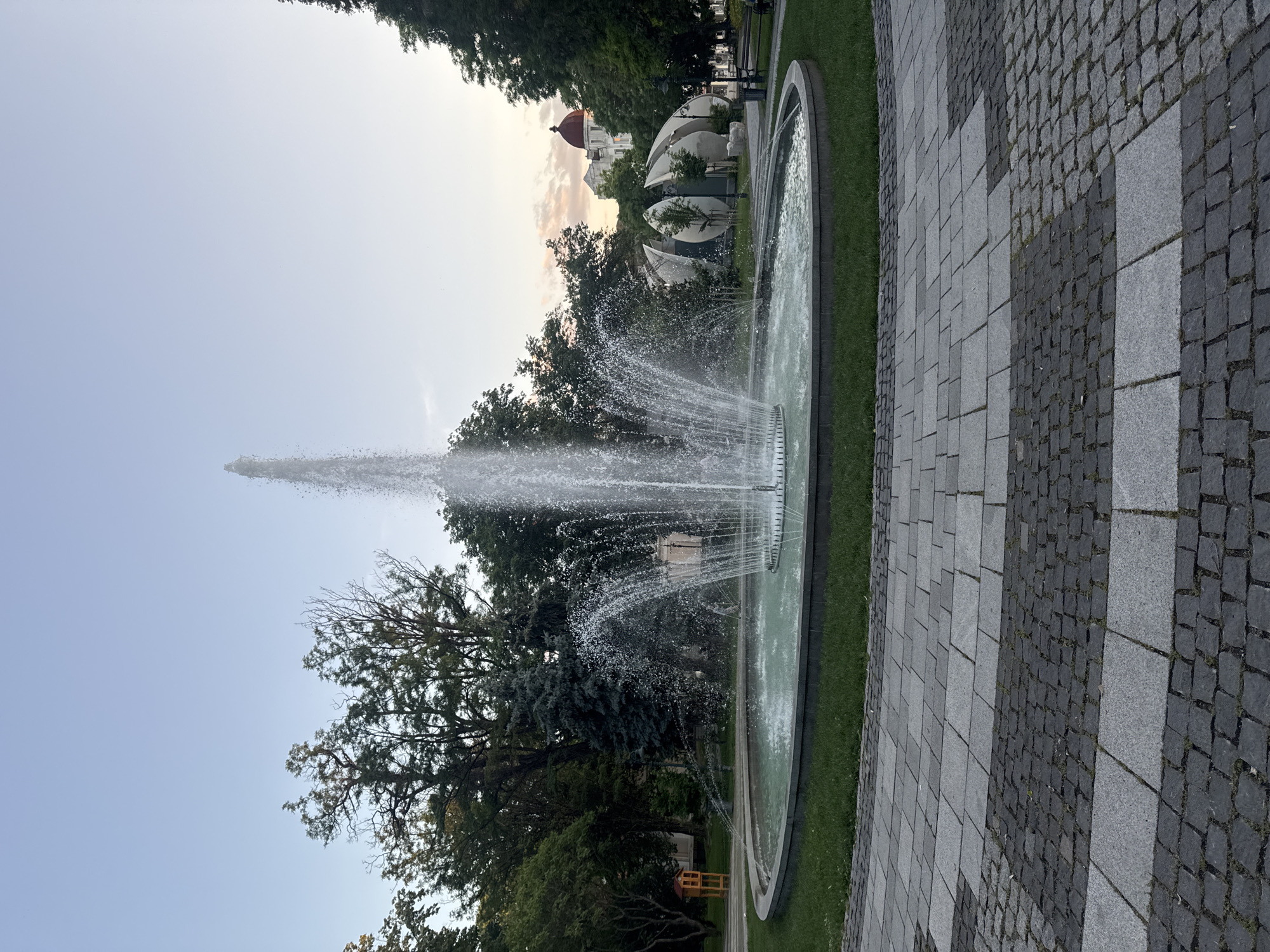 This full-frame, eye-level photograph captures a public square in Plovdiv, Bulgaria, specifically the area around Dzhumaya Square and the Roman Stadium. The central subject is a large, circular fountain with a light turquoise water basin. A tall, powerful stream of water projects vertically from the fountain's center, accompanied by multiple smaller, arcing jets around its perimeter, all actively spraying water.
The fountain is encircled by a manicured patch of green grass. Beyond the grass, a paved area forms a path, characterized by a pattern of alternating sections of large, light grey square pavers and smaller, dark grey cobblestones.
The background features a dense collection of trees, including various deciduous trees with full green foliage and at least one distinct blue-green conifer on the left side.
Architectural details include:
* On the right side of the fountain, a modern, multi-segmented structure, resembling opening white petals or shells, is visible. This is the Plovdiv Roman Stadium Visitor Center.
* Behind this modern structure and further to the right, a large, ornate white building with a prominent, red domed roof is clearly visible.
* On the far left, partially obscured by trees, a small, multi-level wooden structure with an orange-brown façade and a red roof can be seen.
* A park bench is partially visible on the right side of the paved path.
The sky overhead is clear and light blue, transitioning to a warmer, soft orange-pink hue near the horizon on the right, indicative of late afternoon or early evening light. No individuals or animals are present in the observable scene.