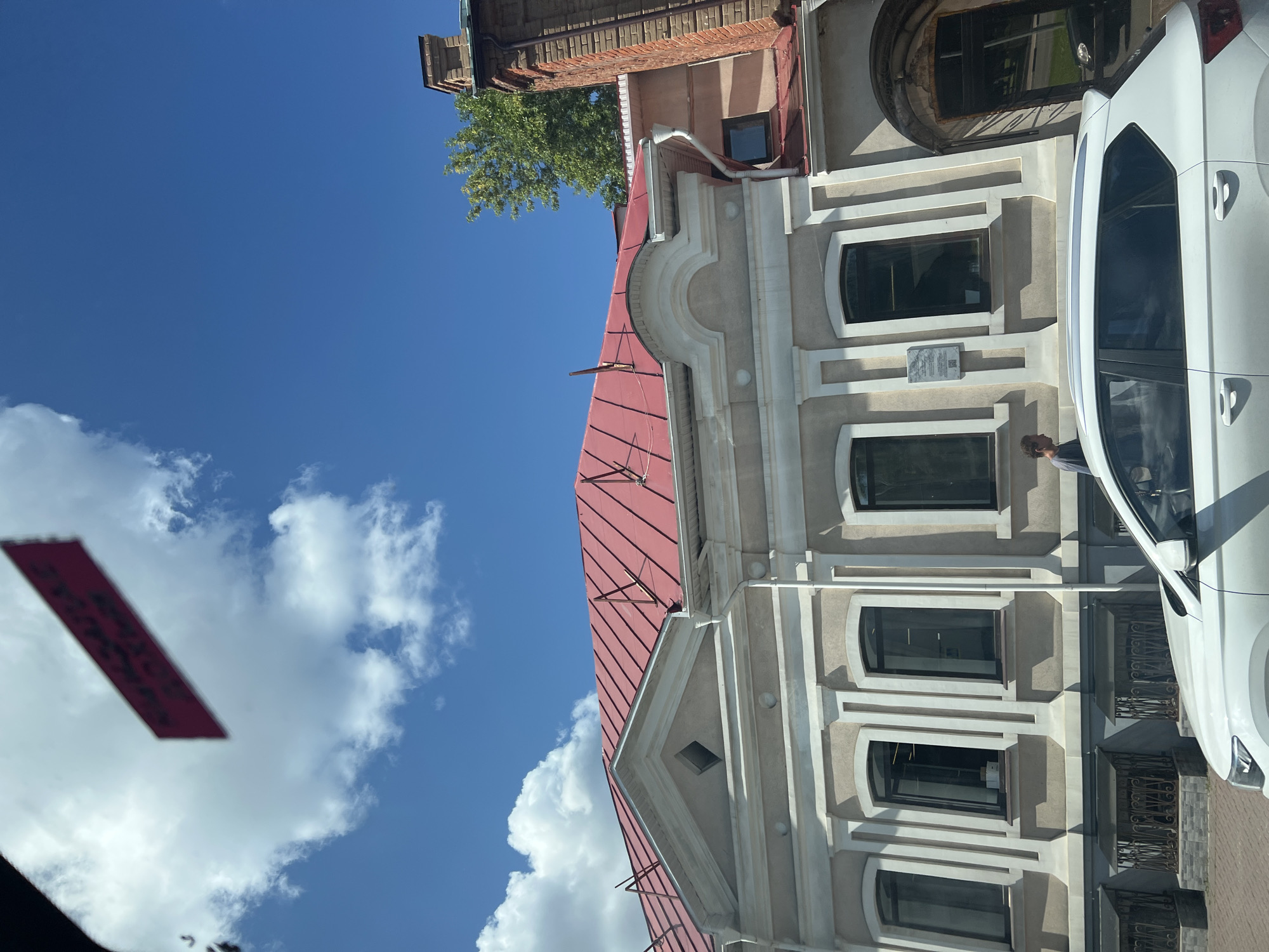 A white SUV is parked on a street in front of a tan building with a red metal roof. The building has several arched windows with dark panes. Above the windows, there is architectural detailing in white. A tree with green leaves grows behind the building, and a brick chimney is visible on the right side. The sky is blue with scattered white clouds. A red rectangular sign with white text is visible in the upper left corner of the image, attached to what appears to be the interior of a vehicle. The sign's text is blurry and difficult to read. A person in a blue shirt is walking on the sidewalk in front of the building. A plaque is mounted on the building wall between two windows. The street is paved with brick or cobblestone.