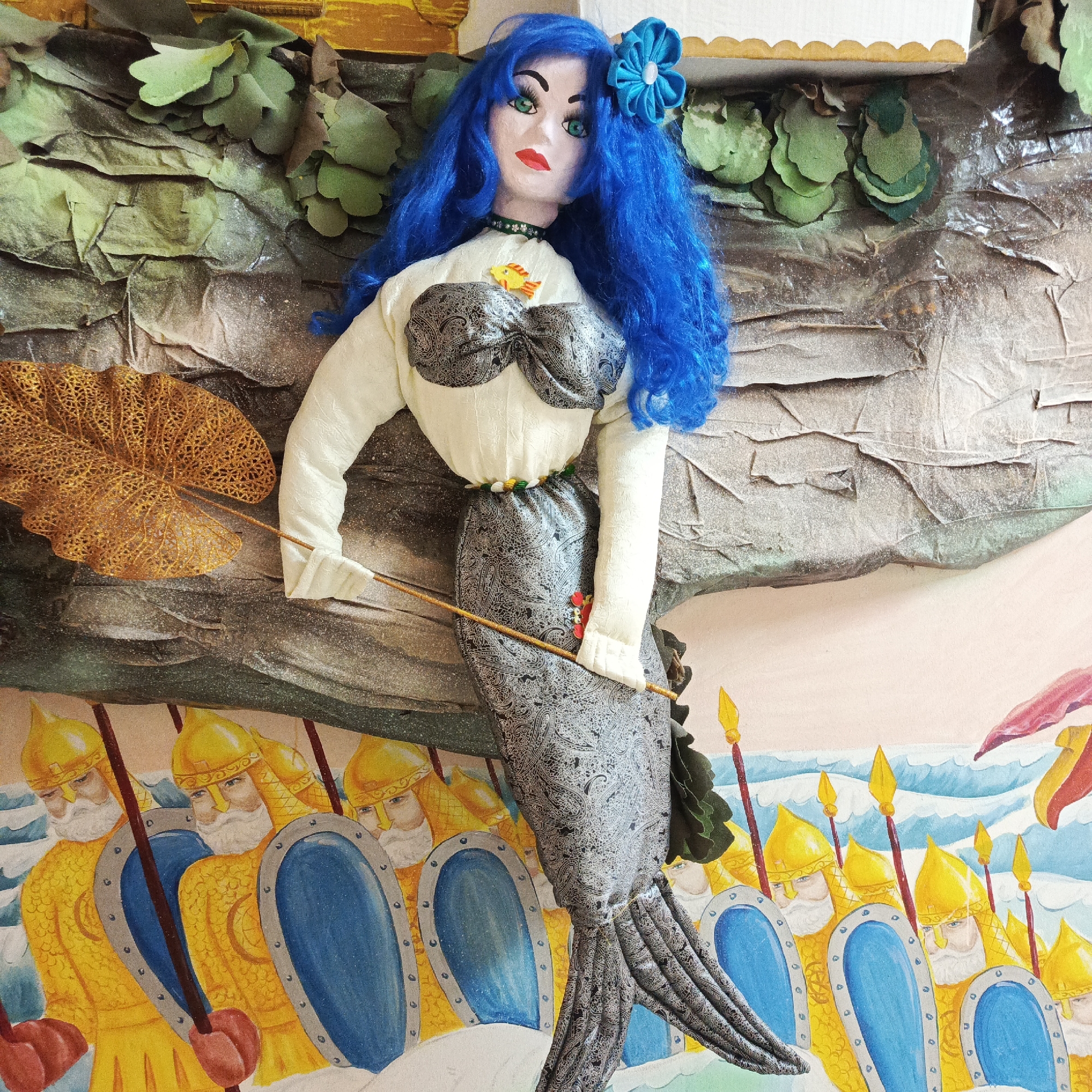 This is a photograph of an artistic display or diorama, likely indoors. The central figure is a handmade doll resembling a mermaid, with blue hair, a white blouse with a black patterned bodice, and a matching gray paisley-patterned tail. She has a blue flower in her hair. She holds a thin brown rod, possibly a spear.
The background appears to be a collage of different materials. Behind the mermaid figure is a textured, possibly paper-based landscape with green foliage. Below the mermaid is a drawing or painting depicting a line of uniformed soldiers with yellow helmets, blue shields, and spears. The scene transitions into an abstract depiction of waves.
The setting is identified as Markovskii, Russia.