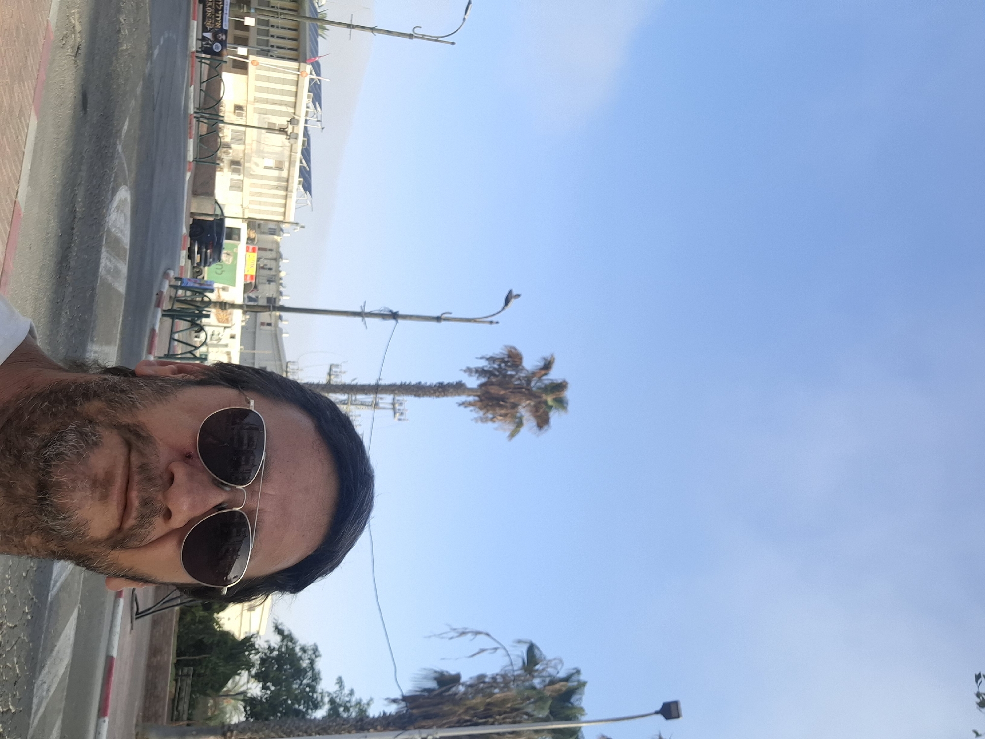 The image shows a man in Yerocham, Israel, wearing sunglasses and a light-colored shirt. He has a beard and short, dark hair. In the background, there is a street with traffic signs, buildings, palm trees, and streetlights. The sky is blue with some clouds. The man is taking a selfie, as evidenced by his position and the framing of the shot. One of the buildings in the background appears to be a school, with solar panels on its roof. There are also advertisements visible on the side of the street.