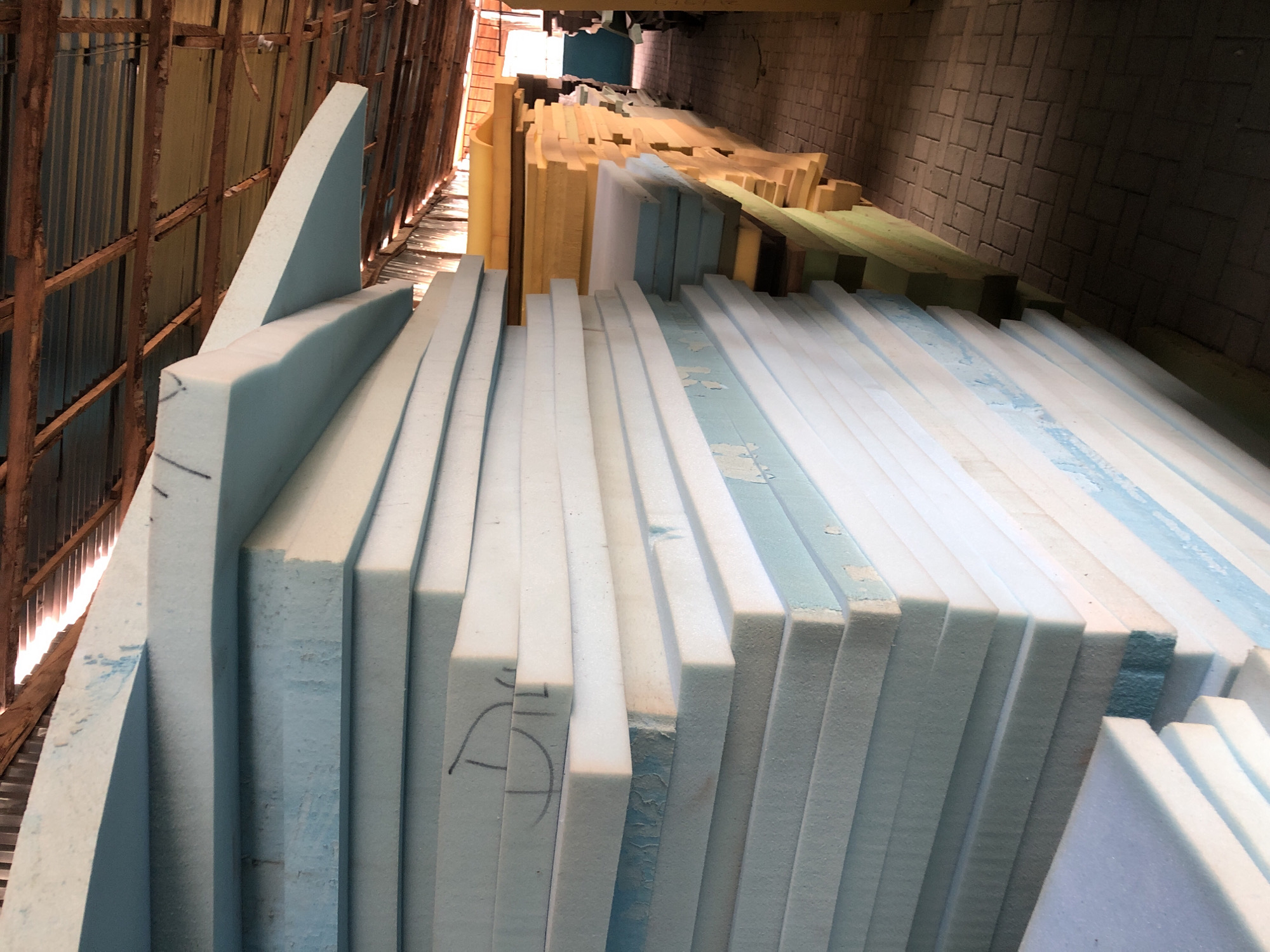 The image shows stacks of foam boards in an industrial setting, likely a storage facility. The majority of the foam is light blue/white and rectangular, arranged vertically. Some boards have markings on them. Deeper in the image are stacks of yellow and green foam. The environment suggests a warehouse or factory, given the corrugated metal walls and ceiling, and concrete block wall in the background. No people are visible. The image does not provide sufficient information to confirm the location is in Ado, Nigeria.