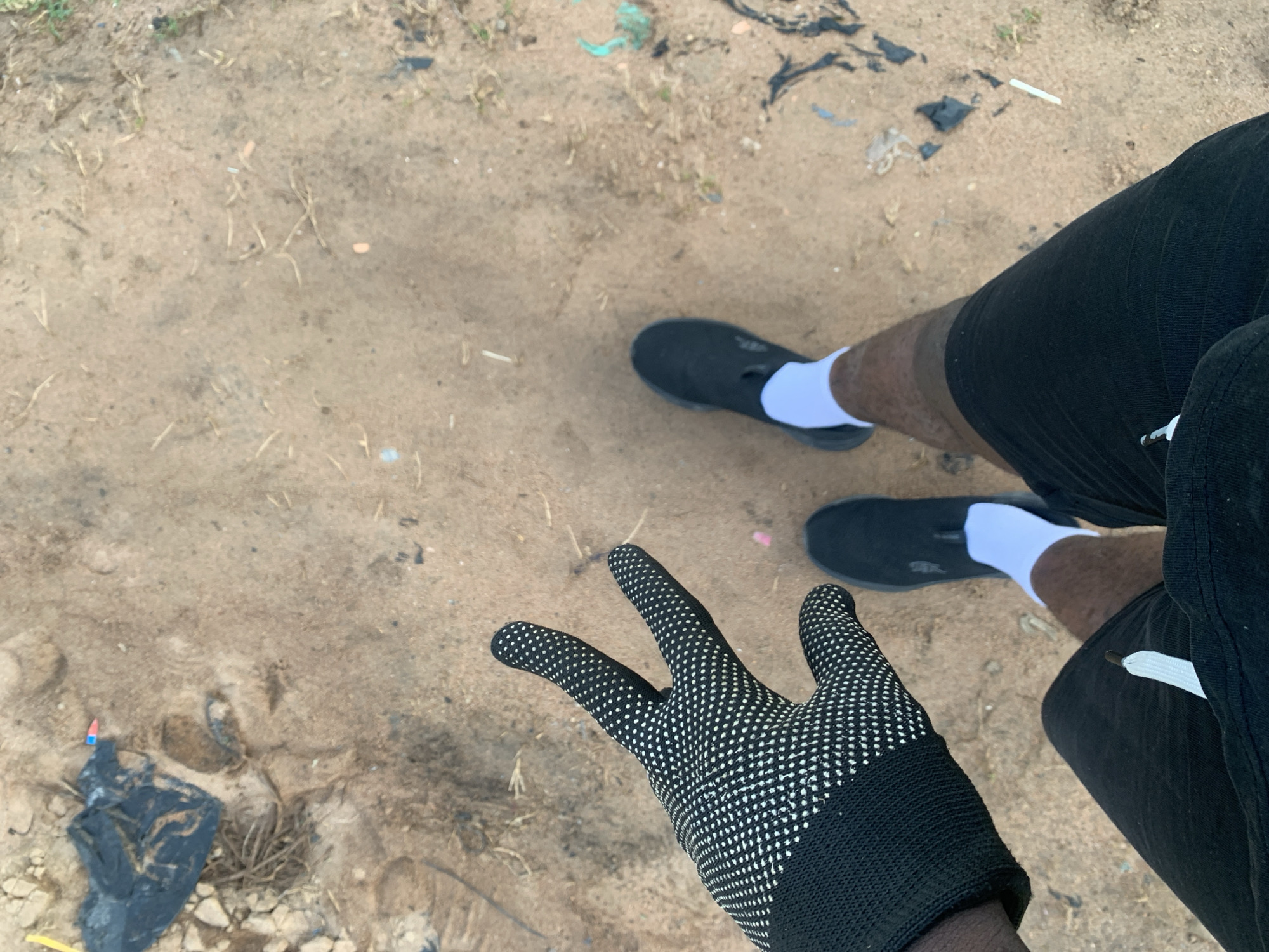 A first-person perspective shot from Jalingo, Nigeria, shows a person's legs and gloved hand. The person is wearing black shorts, white socks, and black slip-on shoes. Their left hand, clad in a black glove with white polka dots, is raised in a peace sign. The ground is a mixture of sand and dirt, with scattered debris including a black plastic bag, small pieces of paper, and dried grass. The lighting suggests an outdoor daytime setting.