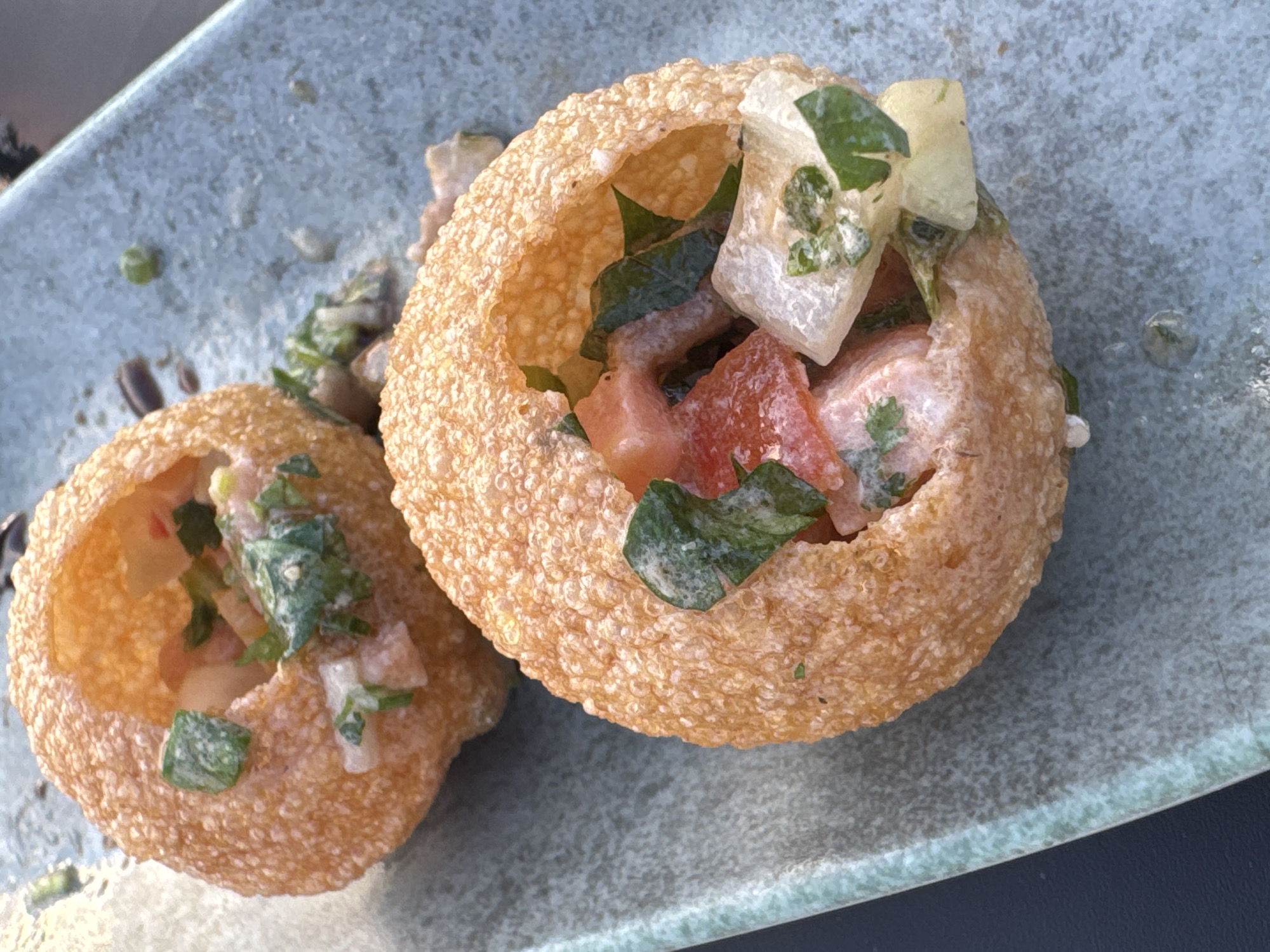 The image displays two golden-brown, spherical, fried pastries, consistent with *pani puri*, presented on a light blue-grey rectangular ceramic plate with a speckled texture. The scene is located in Ness Ziona, Israel. Each pastry is open at the top, revealing an internal filling. This filling consists of diced red components (likely tomatoes), irregularly shaped white or pale-colored pieces (possibly potatoes or onions), and distinct green leafy fragments (consistent with herbs like cilantro or mint). A light-colored, possibly creamy or watery, dressing coats the interior ingredients. The pastry in the upper-left is partially visible, while the one in the lower-right is more centrally positioned. The plate surface shows minor debris, including small green specks and slight liquid traces. A dark, unidentifiable surface serves as a background element on the left and lower-left portions of the frame. No individuals are present in the image.