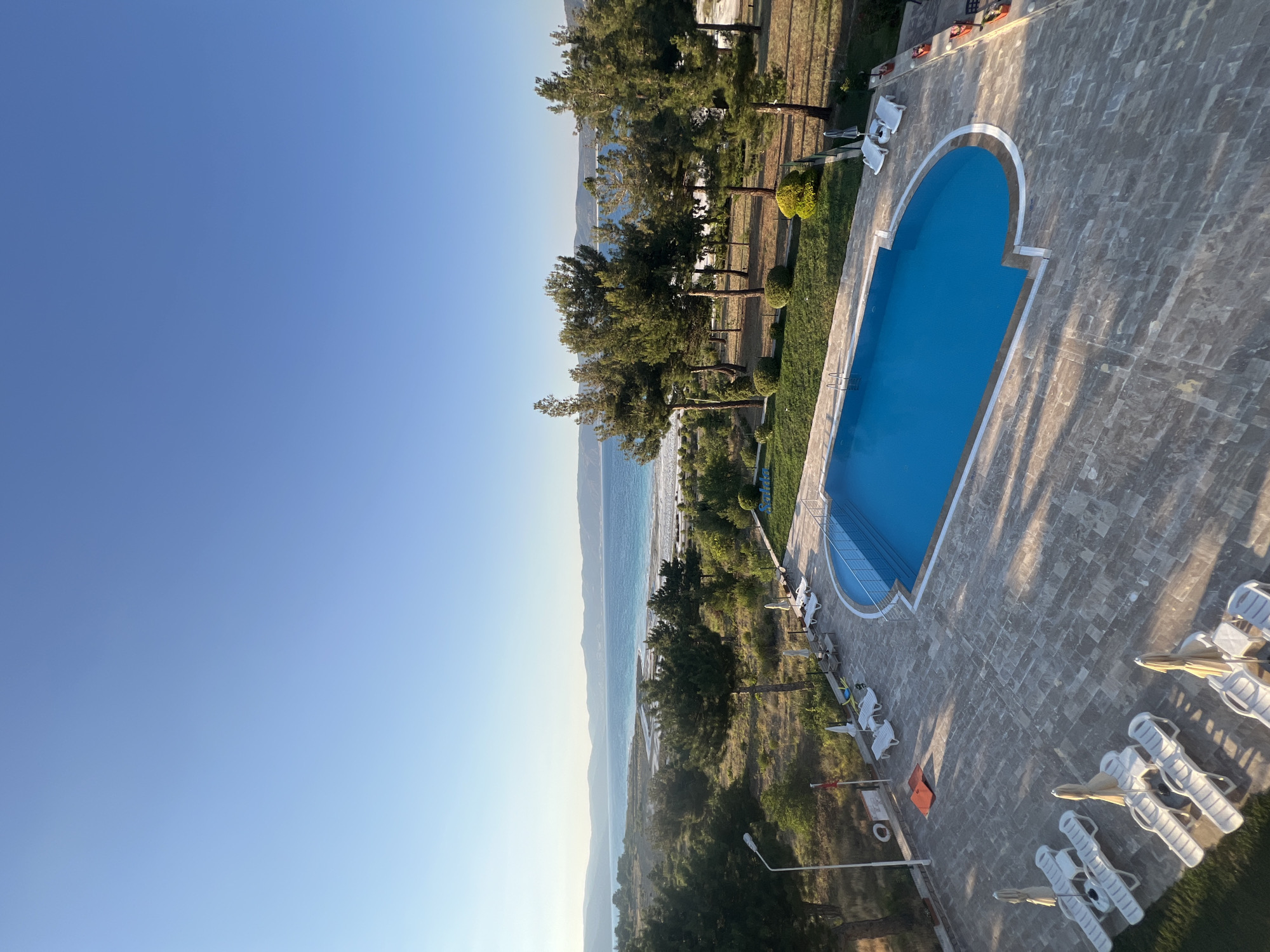 The image shows an outdoor swimming pool at a resort located near the Salda Lake in Yeşilova, Turkey. The pool is rectangular with rounded corners and filled with bright blue water. Lounge chairs and umbrellas are arranged on the paved area surrounding the pool. Beyond the pool area, there are trees and vegetation, with the distinctive white-sand shore of Salda Lake visible in the distance. The lake itself is a vibrant blue, contrasting with the lighter sandy areas along its edge. Mountains form the backdrop, stretching across the horizon under a clear, pale blue sky. The sun is low in the sky, casting long shadows from the trees and structures onto the ground. There are no people visible in the scene.