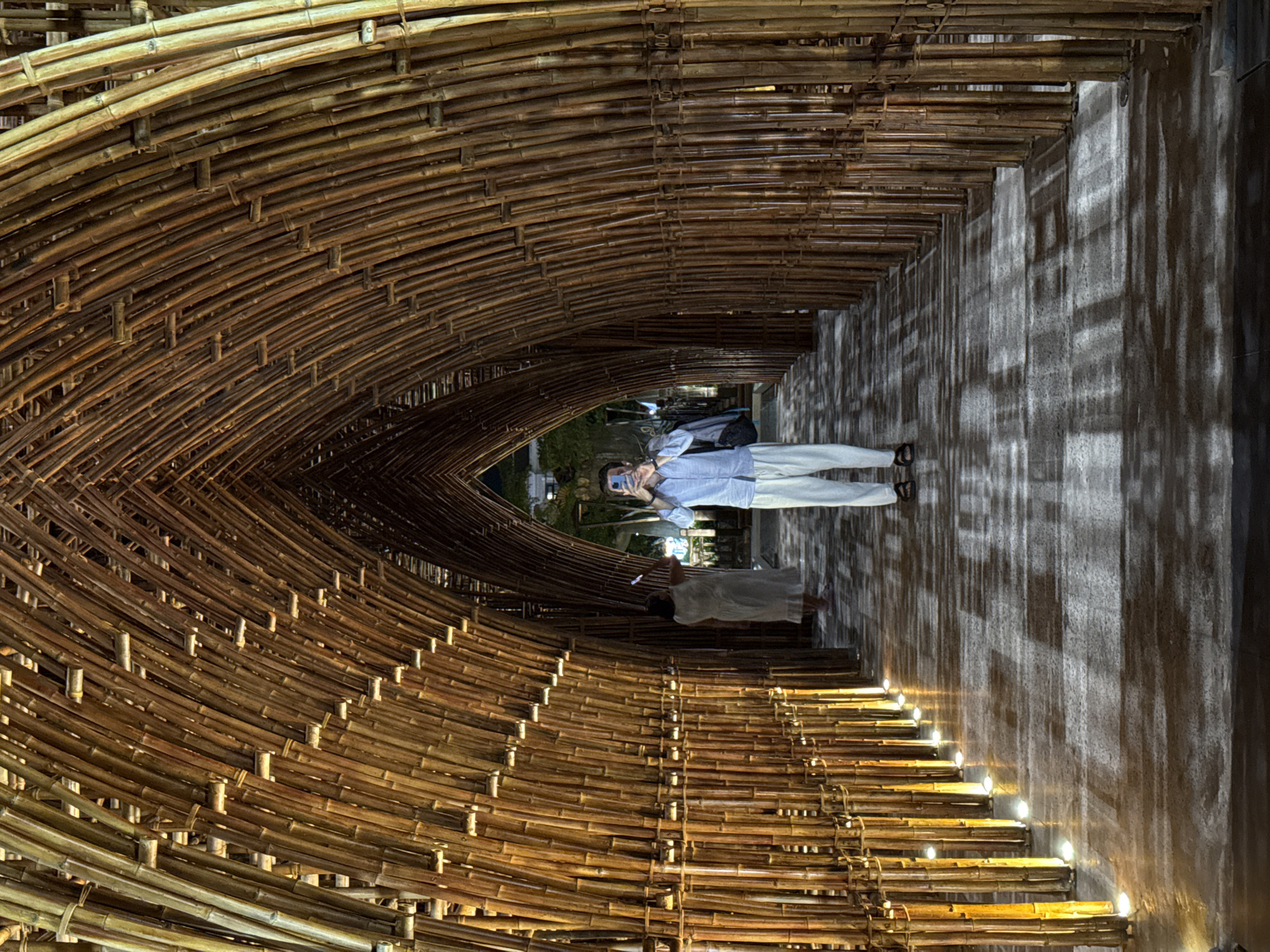 An evening or nighttime indoor shot captures the arched interior of a bamboo tunnel, likely an architectural or artistic installation in Phú Quốc, Vietnam. The tunnel is constructed from numerous bamboo poles, intricately woven and tied together, creating a warm, organic texture that dominates the frame.
Two people are present within the tunnel. In the mid-ground on the right, a person wearing a light blue long-sleeved shirt, light-colored pants, and dark shoes holds up a smartphone, appearing to be taking a photograph or video of the structure ahead. Their face is partially obscured by the phone. Further back and slightly to the left, another person is visible, wearing a light-colored dress, partially obscured by shadows and distance.
Small, warm-toned lights are embedded along the bottom edge of the bamboo structure on the right, casting striking vertical shadows from the bamboo onto the adjacent wall. This wall, appearing to be concrete or a similar light-colored material, reflects the light and shadows, adding depth and visual interest to the scene. The ground beneath the subjects appears to be a flat pathway. The tunnel recedes into the distance, with the intricate bamboo framework creating a strong sense of perspective. The overall ambiance is serene and inviting, highlighting the natural beauty and craftsmanship of the bamboo structure under artificial lighting. There is no visible text in the image.