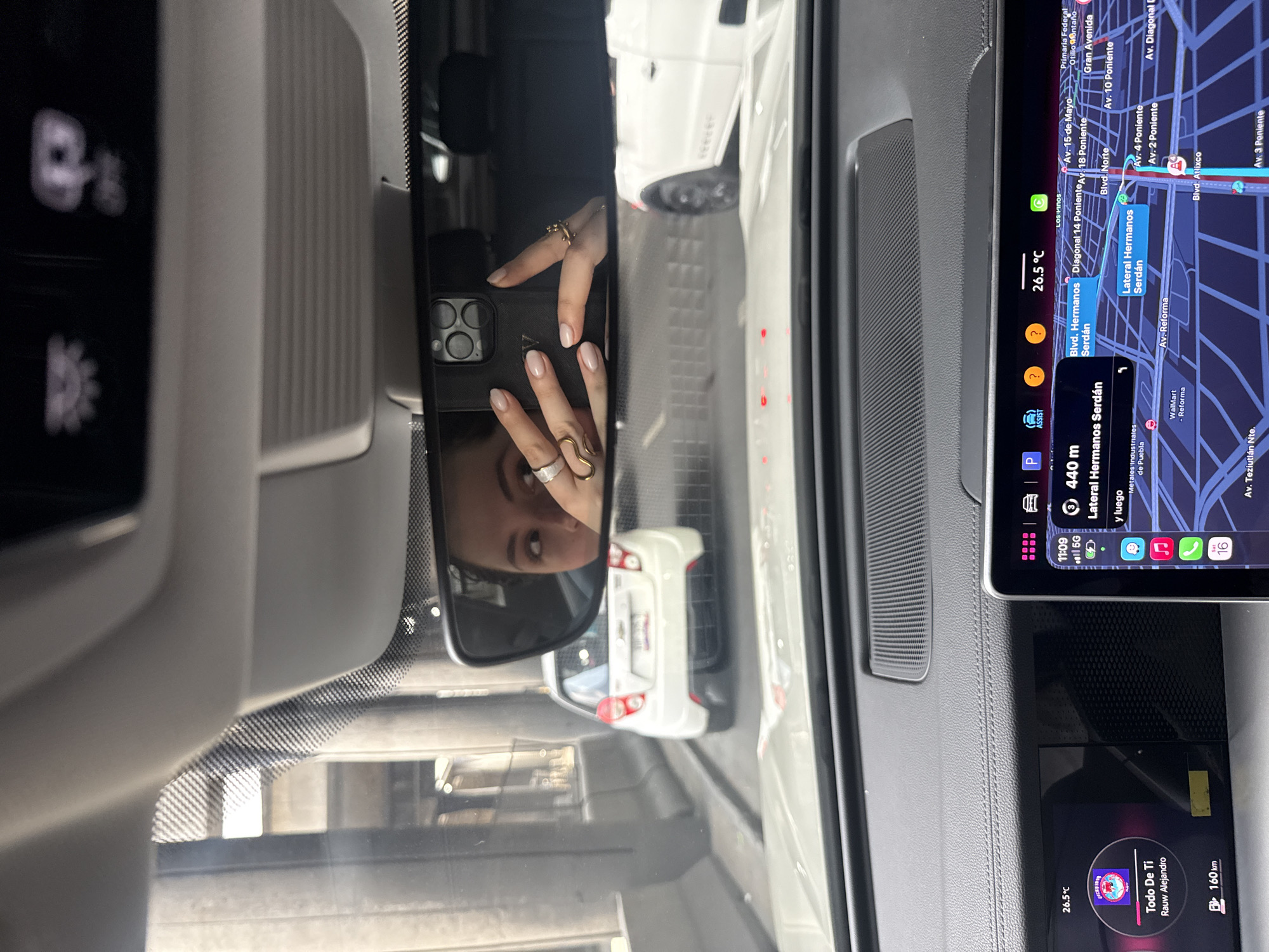 The image shows the interior of a car with a rearview mirror reflecting a person holding a smartphone. The car is located in Puebla, Mexico, as indicated by the GPS navigation screen displaying "Lateral Hermanos Serdán" and other street names specific to that city. The screen also shows a temperature reading of 26.5°C. Below the GPS screen is a secondary screen displaying music information, with the song "Todo De Ti" by Rauw Alejandro being played. The reflection in the rearview mirror reveals the person holding the phone is wearing rings on their fingers. Outside the car, other vehicles are visible.