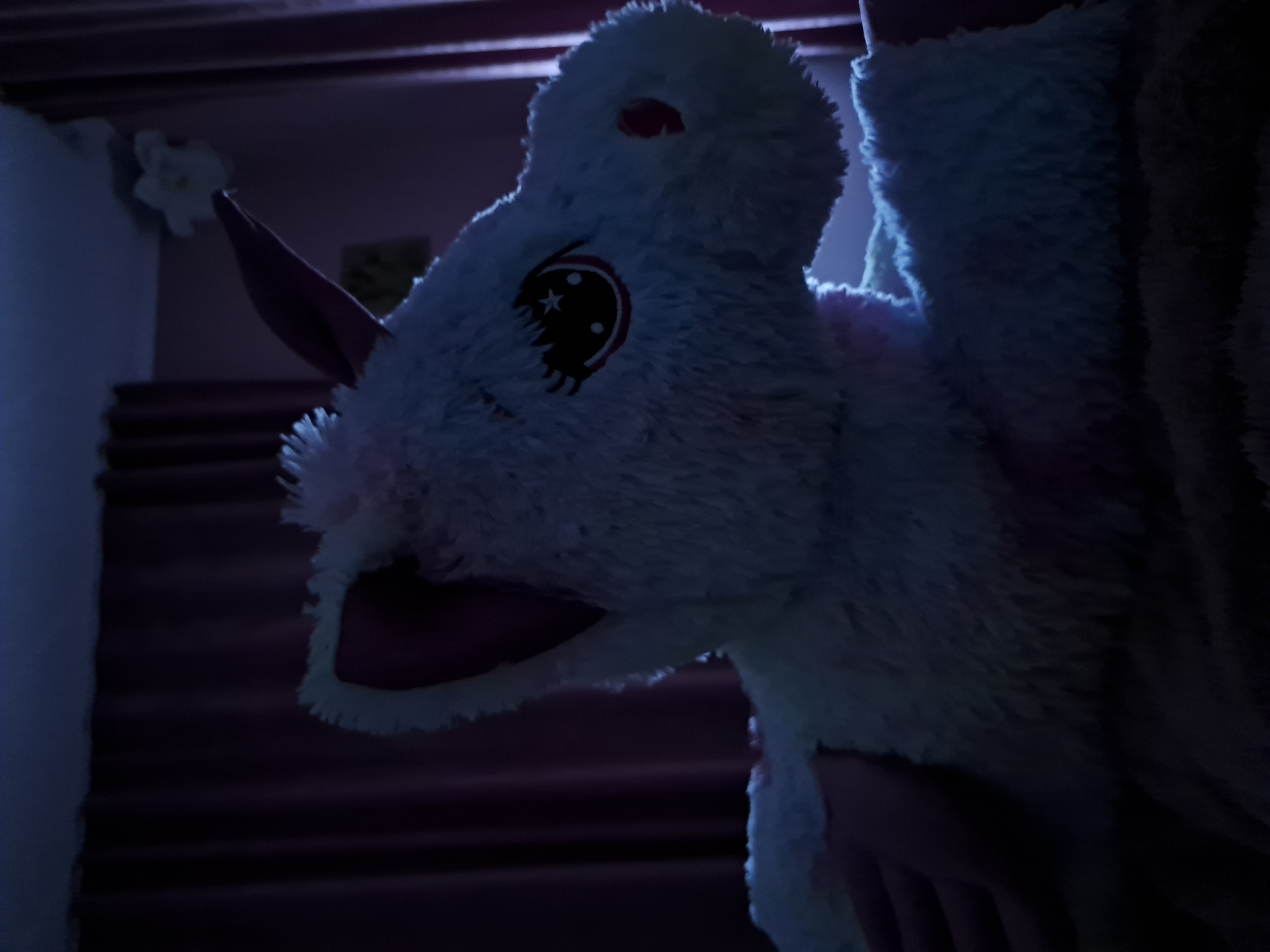 The image captures a close-up of a light-colored, fluffy plush animal head, likely a costume, held by a person. The plush features a prominent black and white eye with a star and jagged design, a pointed ear, and a snout, along with a small red detail on top of its head. The material is distinctly soft and furry.
The setting is indoors and dimly lit, suggesting nighttime or a low-light environment. In the background, a dark staircase with multiple treads is visible, receding upwards and to the left. A light-colored wall with a subtle white floral decoration is also present on the left side of the frame. The person holding or wearing the plush costume is largely out of frame, with only a hand and shoulder faintly visible, interacting with the fluffy material. No text is visible in the image. The overall mood is subdued due to the low light conditions.