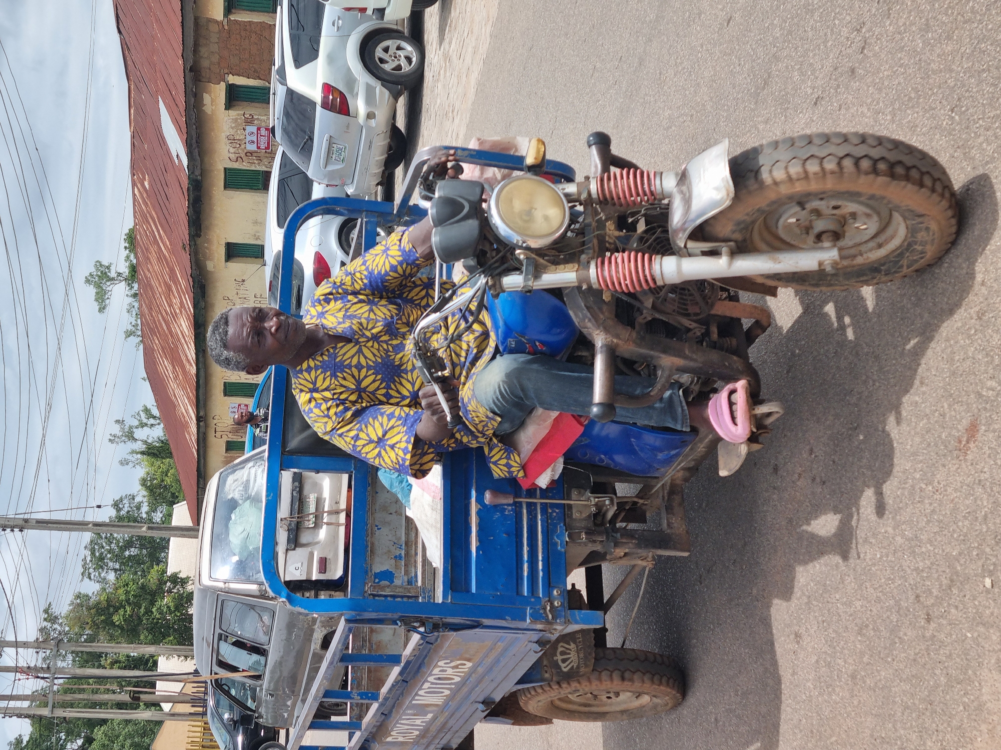 A grey-haired adult male, likely in his 50s or 60s, operates a blue three-wheeled cargo motorcycle (a tricycle) on a paved street under a partly cloudy sky in what is identified as the city of Jos, Nigeria. He wears a blue and yellow patterned short-sleeved shirt and blue jeans, with a pink slide sandal on his left foot which rests on a pedal. His hands are on the handlebars, and he looks slightly towards the viewer's right.
The blue tricycle features "ROYAL MOTORS" stenciled in white on its cargo bed and a "ROYAL MOTORCYCLE" crown logo on the rear wheel arch. The vehicle shows wear, including chipped paint and worn tires. The front wheel has a silver fender and red-coiled suspension springs.
In the mid-ground, a silver passenger van, appearing to be loaded, is positioned behind the tricycle. To its right, a silver SUV with the Nigerian license plate "JGY-605L" is visible. Other vehicles are partially visible further in the background.
The background includes a weathered, single-story building with a rusted, reddish-brown corrugated metal roof and light-colored, possibly tan or yellow, walls. It features multiple green-framed windows. Two white signs with red lettering are affixed to the building: one reads "STOP URINATING HERE," and the other similarly states "STOP URINATING HERE" with a red no-urinating symbol and additional obscured text below, likely "REFUSE AND..."
Utility poles and numerous overhead wires crisscross the sky. Trees with green foliage are visible behind the buildings. Strong shadows cast on the asphalt pavement indicate a sunny environment. A second individual, a dark-skinned male in a striped shirt, is partially visible in the far background behind the silver van and to the right of the tricycle's bed.