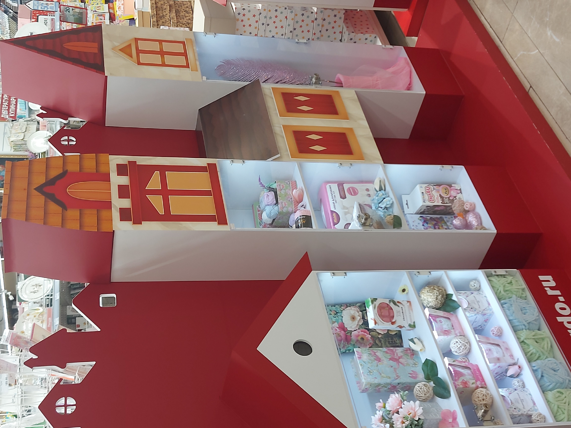 The image shows a multi-tiered product display in a store. The display is designed to resemble a series of houses, stacked on top of one another. Each "house" features a white interior containing various goods, including decorative items, crafting supplies, and small gifts. The exteriors of the "houses" are red and beige, with painted details such as windows and roofs. The display is set against a red background and is positioned on a light-colored tile floor. The objects in the display are arranged in sections. The photograph was most likely taken at a store located in the city of Saint Petersburg, Russia, based on the URL "do.ru" in the bottom right corner.