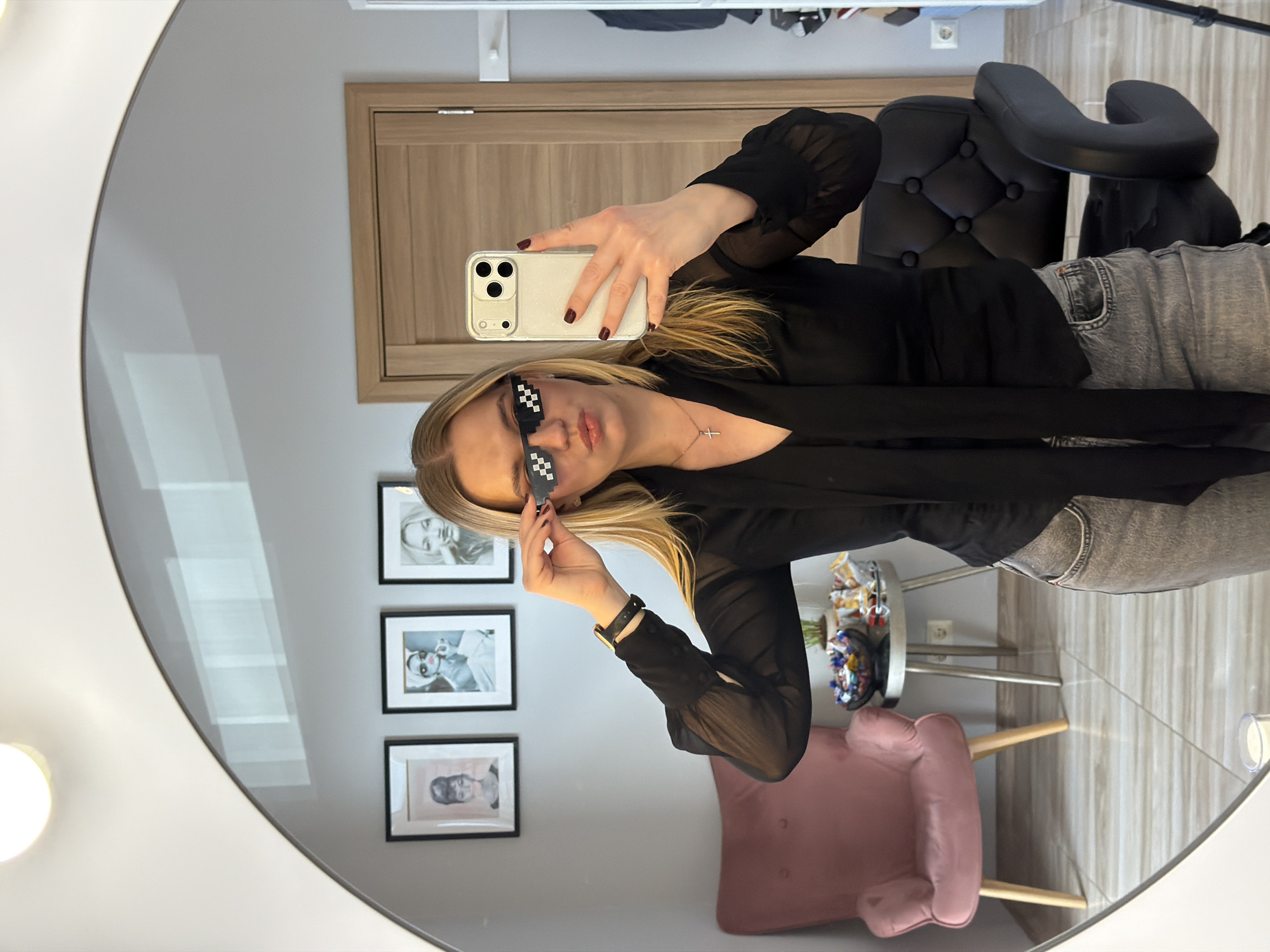 A woman is taking a selfie in a mirror. She is wearing black pixel sunglasses, a black top, and gray jeans. She has blond hair and is wearing a cross necklace. The location appears to be indoors, possibly in Riga, Latvia, due to the design and furnishings. In the background, there are framed pictures on the wall, a pink armchair, and a black chair. There is also a round table with assorted items on it. The mirror is round and reflects part of the room.