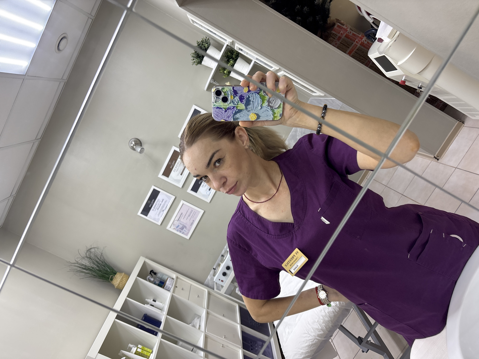 A woman is taking a mirror selfie in what appears to be a salon or medical office. She is wearing a purple scrub top and has blonde hair pulled back. She is holding up a phone with a floral case to take the picture.
In the background, there are shelves with various bottles and jars, suggesting a retail or treatment space. There are also framed certificates on the wall, and a plant in a woven basket. The room is brightly lit, possibly with fluorescent ceiling lights. The overall impression is one of a professional, clean environment. The woman's expression is neutral, and she is looking directly at the camera. The time of day is not discernible from the image. The location cue "Medovoye, Russia" is provided, but no specific visual elements in the image directly indicate this location.
There is visible text on a yellow nametag pinned to her scrub top, which appears to read "YUIIA".