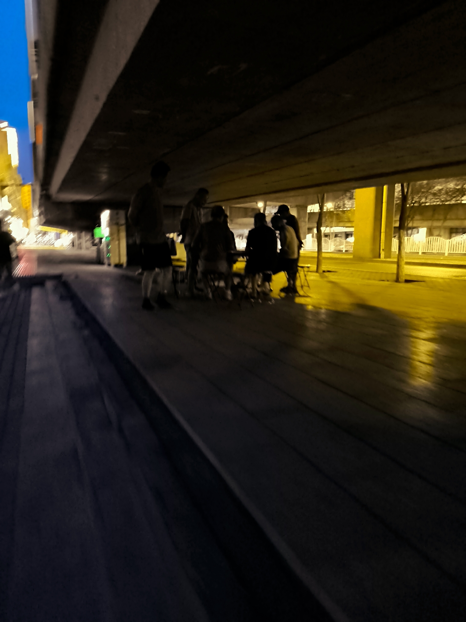A group of people are gathered under a bridge at night. The lighting is dim, with a strong yellow cast from streetlights. The ground appears to be made of dark wood planks. The people are silhouetted, making it difficult to discern details. One person in the foreground, on the left, is standing and facing away from the camera. Further back, a group of several individuals are clustered together, some seated, possibly around a table or at chairs. The background shows more urban elements, including buildings, streetlights, and a fence. The overall atmosphere is subdued and possibly informal, given the setting.