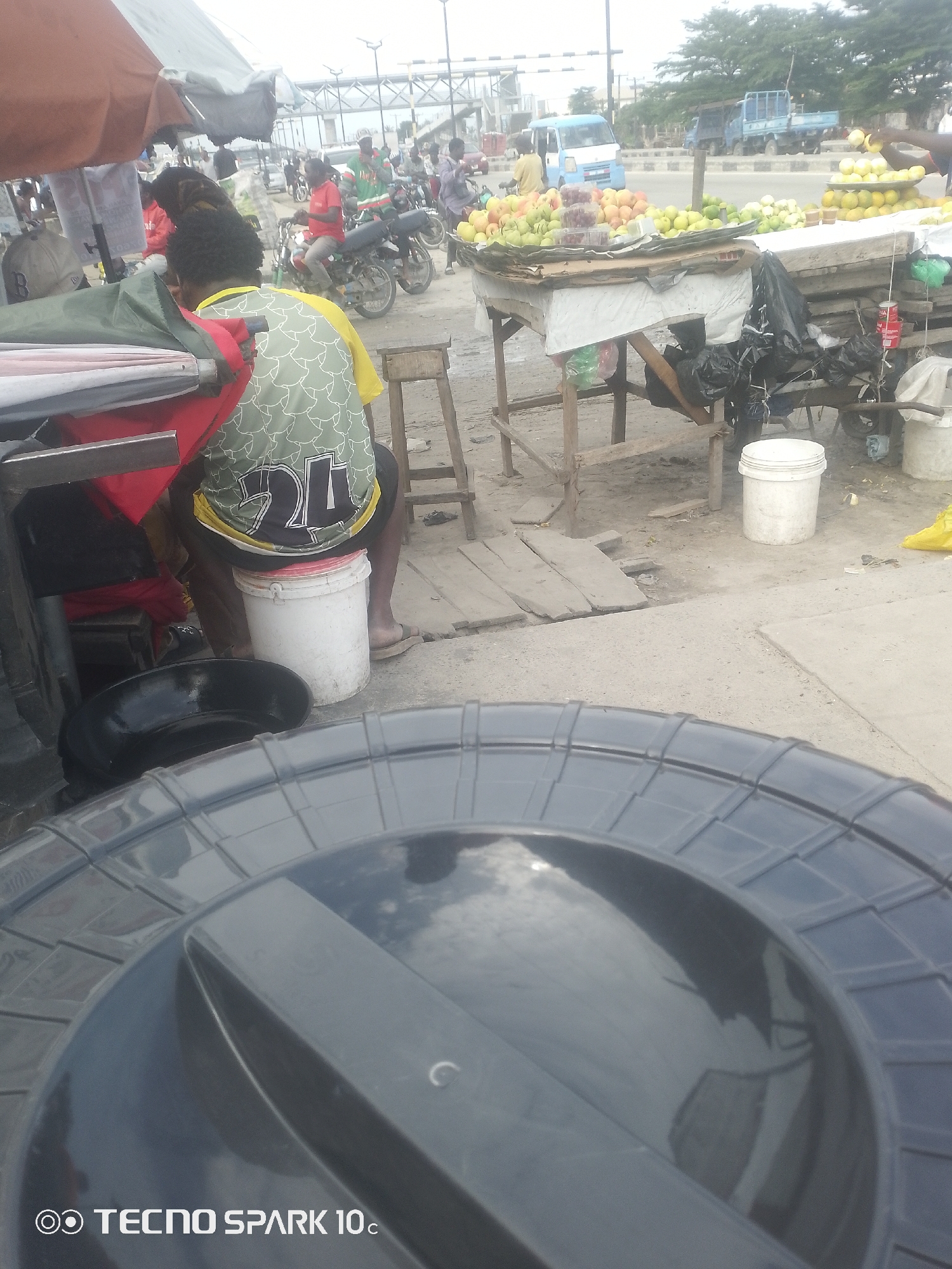 This image depicts an outdoor market scene in Adesanya, Nigeria. The foreground is dominated by the lid of a large, dark plastic container, possibly a water tank, reflecting the sky and surrounding environment.
In the mid-ground, a person wearing a green and yellow patterned shirt with the number "24" is seated on a white bucket. They are facing away from the camera, and their legs are visible, wearing sandals. Behind them, part of a stall with a red cloth and other items is seen.
Further back, several other individuals are present, some appearing to be vendors or shoppers. A person in a red shirt is seen on a motorcycle, and others are visible around stalls laden with fruits, including what appear to be apples, grapes, and lemons. A blue truck and other vehicles are in the background, suggesting a busy roadside market.
The setting appears to be a market located alongside a road, with various stalls and temporary structures. The ground is dusty and uneven, with some wooden planks laid out. The weather appears to be overcast or hazy, and the time of day is likely daytime. The overall impression is that of a lively, everyday market in Nigeria.
The visible text in the image is "TECNO SPARK 10c" located at the bottom left corner.