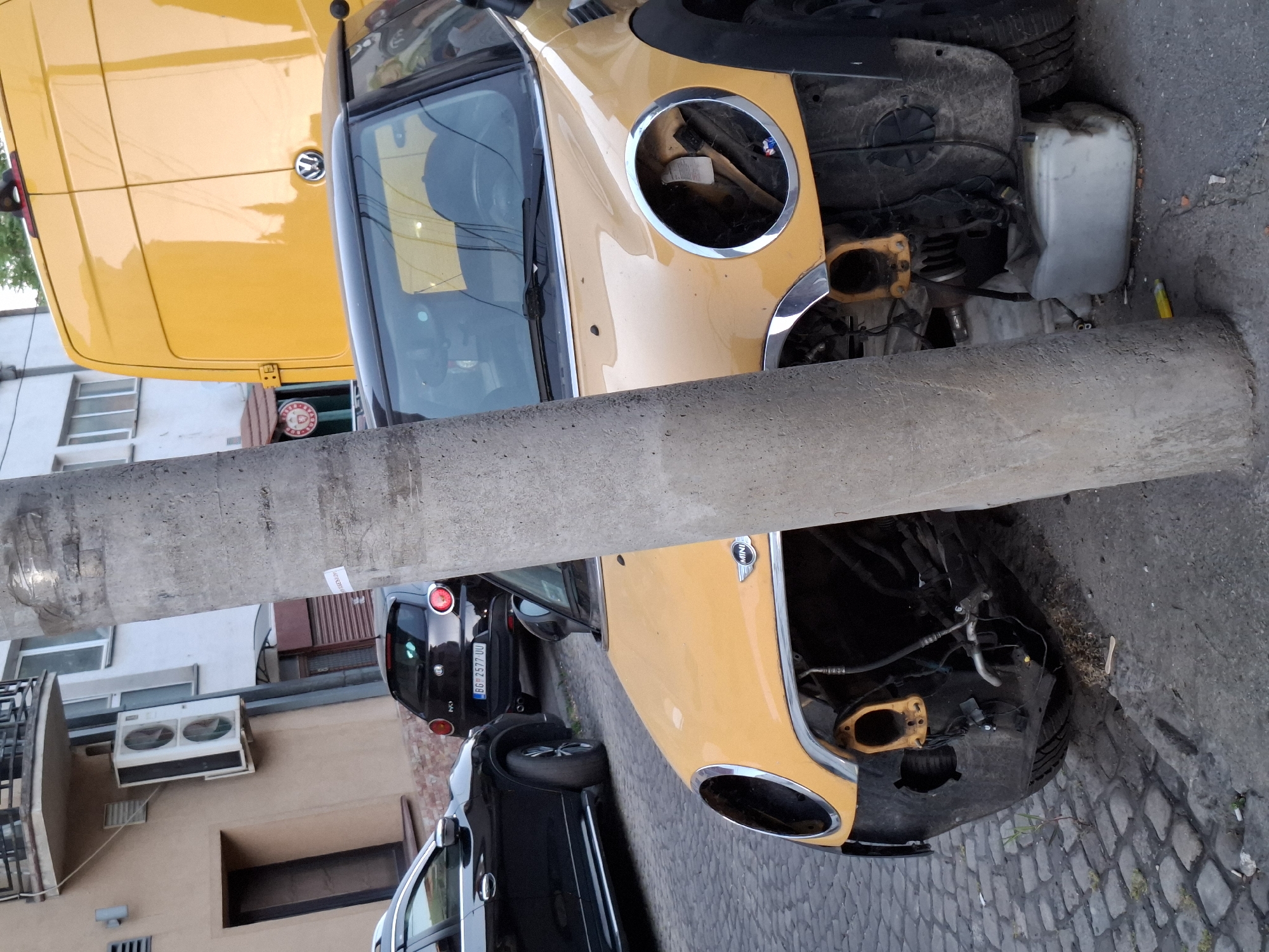 The image depicts a motor vehicle accident in an urban environment, likely in Zemun Urban Municipality, Serbia. A yellow Mini Cooper is severely damaged, with the front end missing and impacting a concrete utility pole. The vehicle is wedged between the pole and the curb of a cobblestone street. A yellow Volkswagen van is parked to the left. There are other parked vehicles nearby, and the license plate "BG-2577-UU" is visible on a black hatchback. The background shows a building with air conditioning units. There are no people visible.