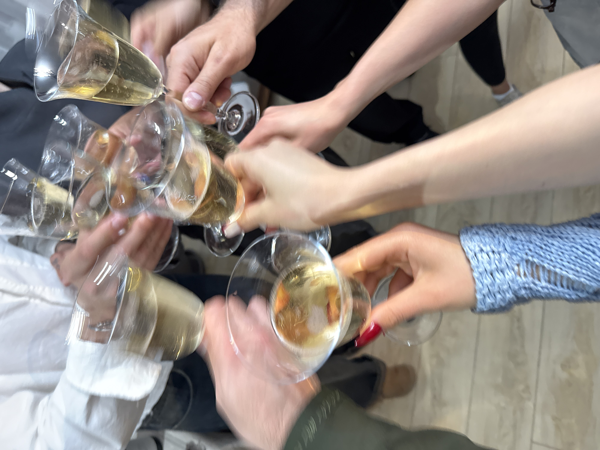 The image shows a group of people toasting with glasses of a pale yellow beverage, likely champagne or sparkling wine. The shot is taken from above, showing multiple hands holding the glasses, which are clinking together. People are wearing various attire including a white collared shirt, black clothing, a blue knitted sweater, and an olive green jacket. The location is indicated to be in Yerevan, Armenia, although there are no specific landmarks or identifiers visible in the image to confirm this.
