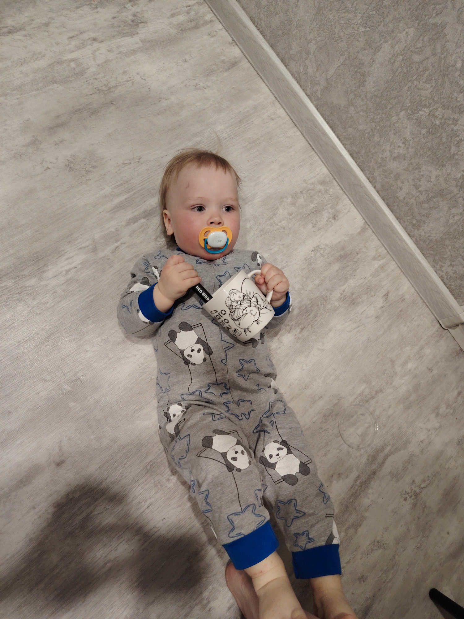 A baby is lying on a grey, wood-look laminate floor in a room in Chaikovsky, Russia. The baby has a light complexion, blonde hair, and a pacifier in their mouth. They are wearing grey pajamas patterned with pandas and blue stars, with blue cuffs. The baby is holding a white mug with cartoon drawings on it. A white baseboard runs along the edge of the floor, next to a grey wall. A bubble is visible on the floor near the baby's feet. The photo is taken from an overhead perspective.