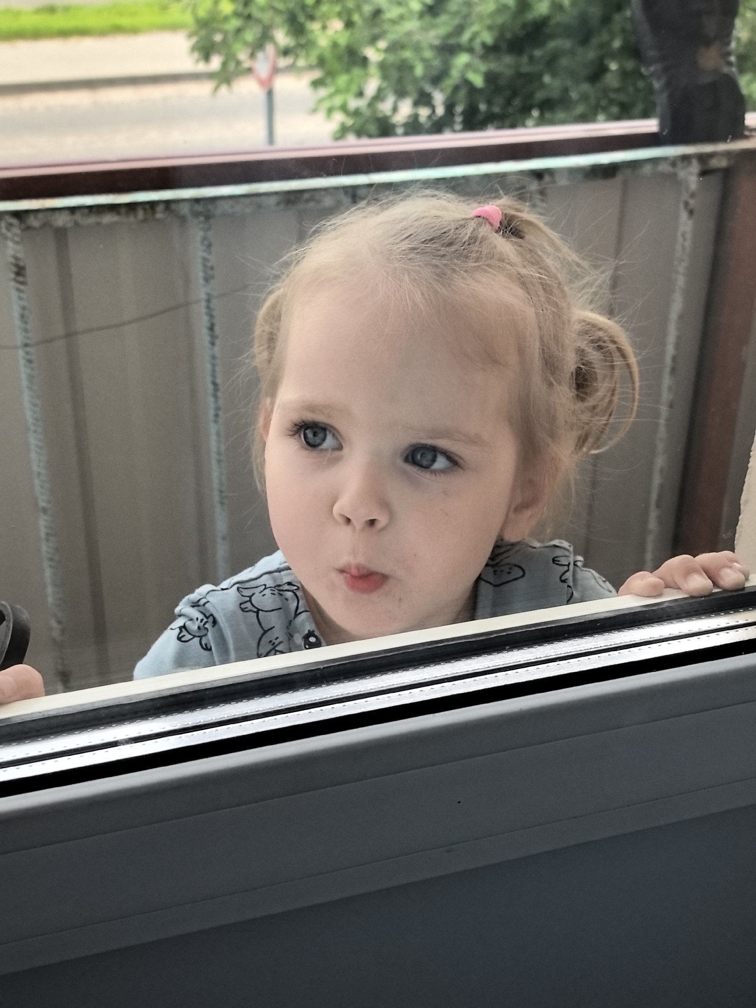 A young girl with blonde hair pulled into two pigtails, one secured with a pink scrunchie, looks curiously out of a window. She is wearing a light blue t-shirt with a pattern of small, outlined animals. Her mouth is slightly pursed, and her blue eyes are wide, suggesting a moment of intense observation. Her hands are resting on the window frame, with her fingers visible on the right side of the frame.
The setting appears to be indoors, looking out onto an outdoor scene. Through the window, blurred greenery, trees, and a street with a red and white traffic sign are visible, indicating it is daytime and likely outdoors. The window frame is a modern design, with a grey lower section and a thinner white and black frame visible at the top. The overall lighting suggests it is a bright day. The girl's expression conveys a sense of innocent curiosity and engagement with the world outside.