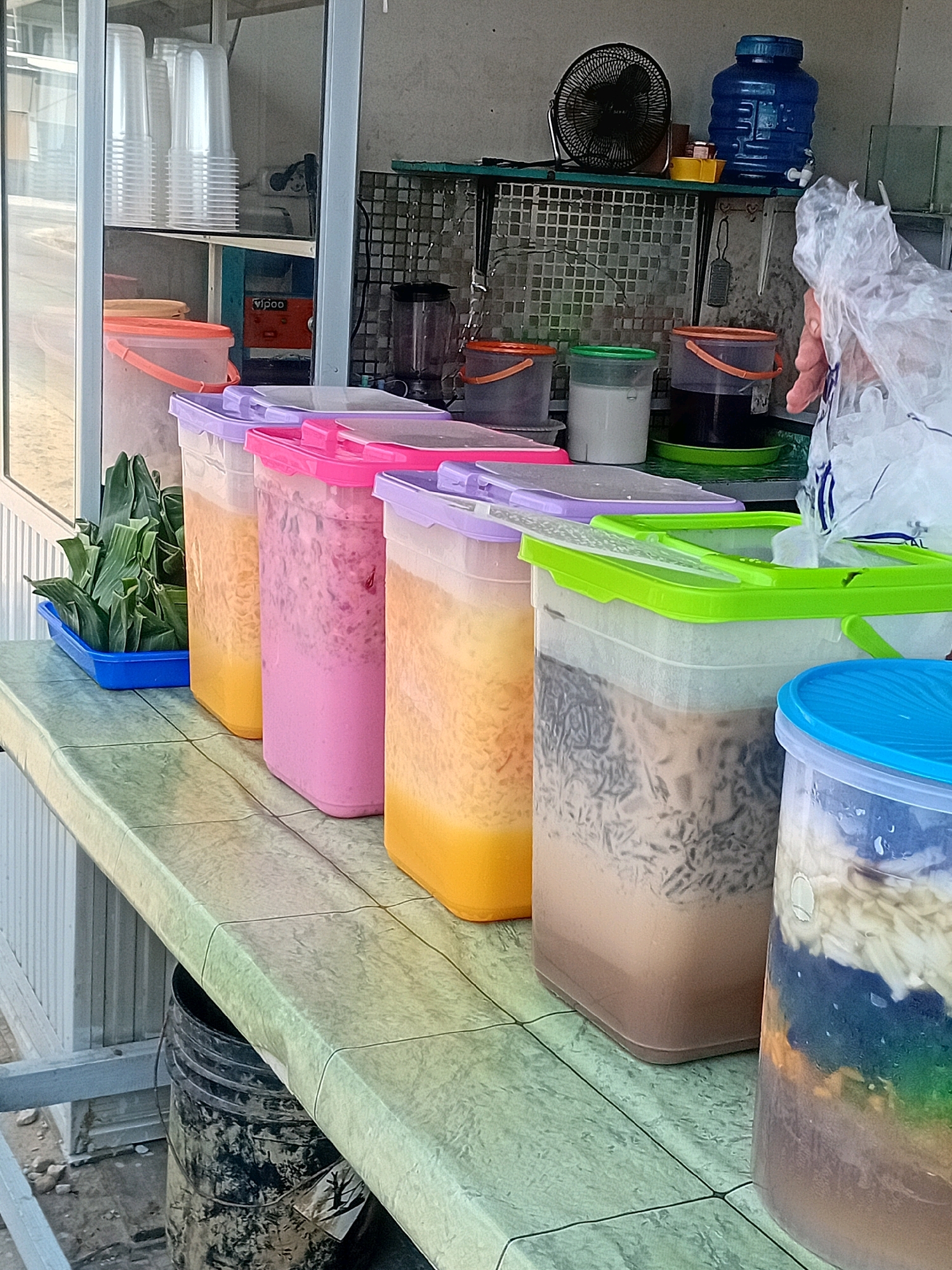 A row of large, clear plastic drink containers with brightly colored lids sits on a speckled green countertop, presumably at a street-side stall in Sungai Meriam, Indonesia. The containers hold various colorful beverages and what appear to be ingredients for desserts or drinks. From left to right, the first visible container has a pink lid and contains a pale pink liquid with flecks of a darker shade, suggesting it might be a fruit-flavored drink. Next, a container with a yellow lid holds a lighter yellow liquid. Following that is a container with a light purple lid containing a beverage with a milky brown base and darker, noodle-like ingredients. The furthest visible container on the right has a bright blue lid and holds layers of colorful ingredients, including what looks like shredded coconut, blue and green jellies, and other unidentifiable components. In front of the drink containers, a blue plastic tray holds a bundle of green, leafy vegetables, possibly ingredients for local cuisine. To the left of the containers, a black plastic bucket with a weathered appearance is visible under the countertop. In the background, a shelf holds stacks of clear plastic cups, a blender, several small plastic containers with lids, a black desk fan, and a large blue water jug. A hand holding a plastic bag filled with ice is reaching into the container with the green lid, indicating recent preparation or serving. The overall setting appears to be an outdoor food or drink stall.