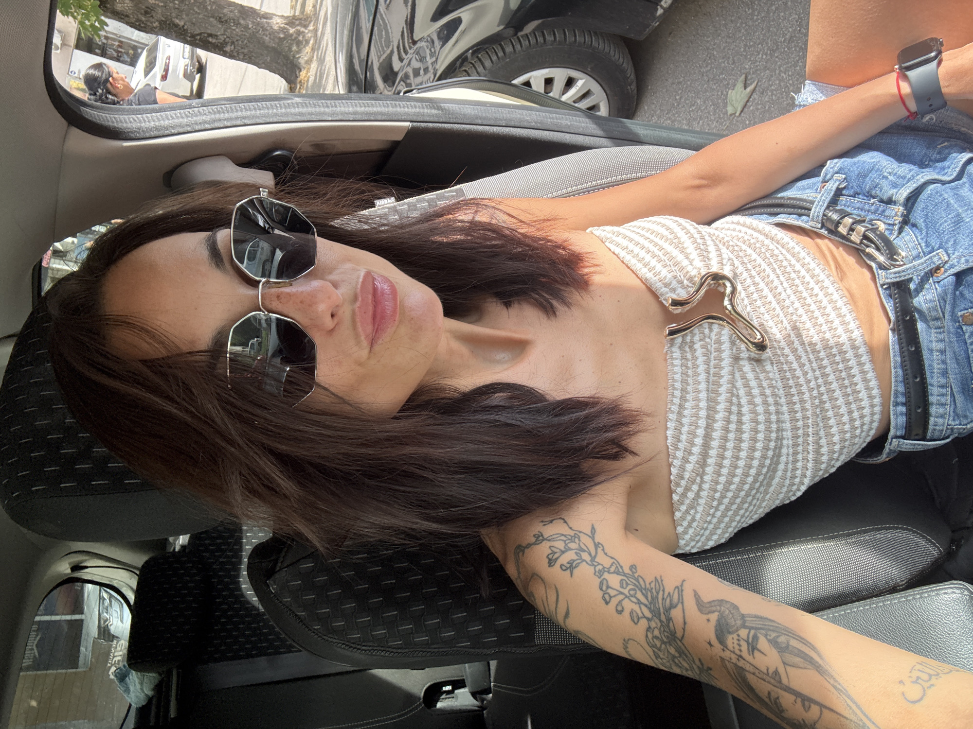 A woman with long, dark hair, wearing sunglasses, a strapless top, and denim shorts, is seated in the passenger seat of a car. Her left arm is visible, showcasing tattoos. A gold-colored accessory is attached to the front of her top. In the background, through the car window, a person is standing near a white car parked on a street, with a tree and buildings visible. The scene appears to be in the city of Pleven, Bulgaria, suggesting an urban environment.