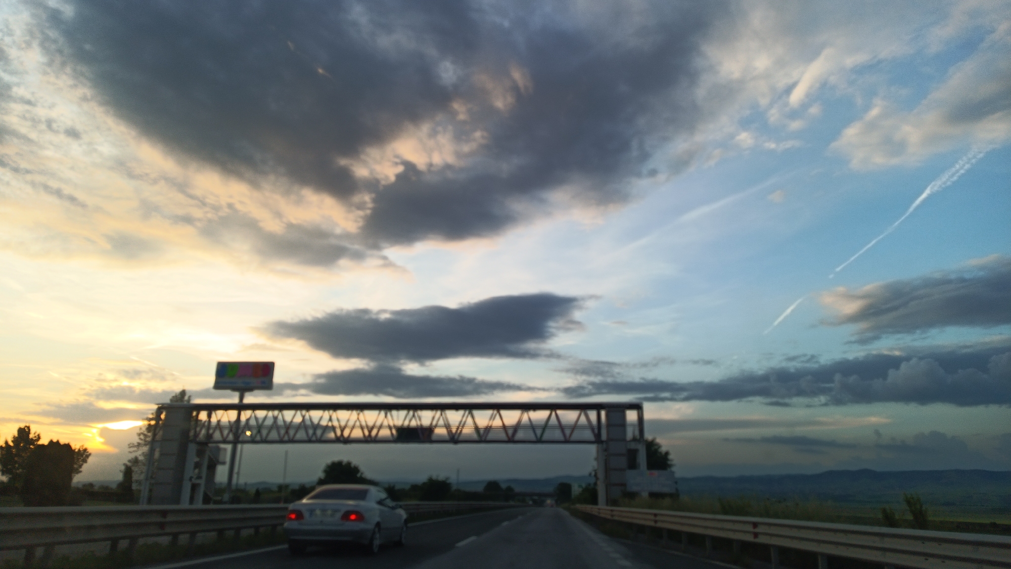 The image depicts a view from a moving vehicle on a highway. A silver car is visible in the adjacent lane. An overhead gantry structure spans the highway, with a sign atop it. The sky above is filled with clouds, with the sun appearing to set on the left horizon. The environment appears to be a rural area with distant hills. The location cannot be determined from the image.