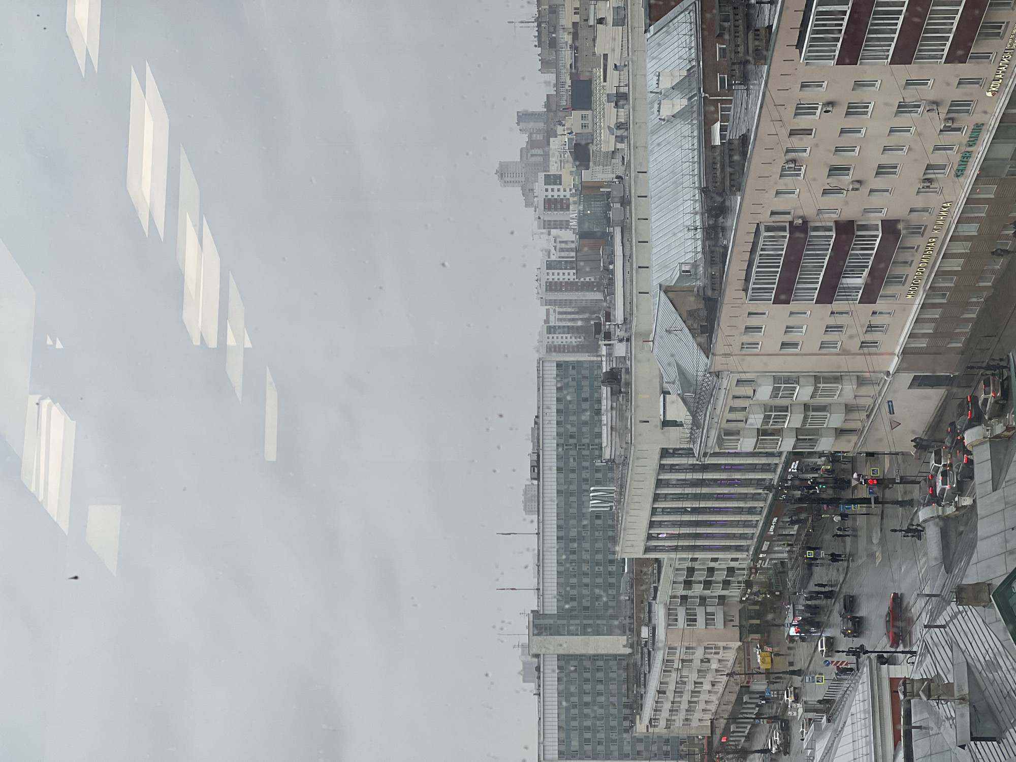 View from a high vantage point in Perm, Russia, showing a cityscape on a rainy day. Buildings, streets, and vehicles are visible. People are walking on sidewalks. There is a building in Cyrillic, possibly an office or clinic. The sky is overcast and the image is partially obscured by raindrops on the window.