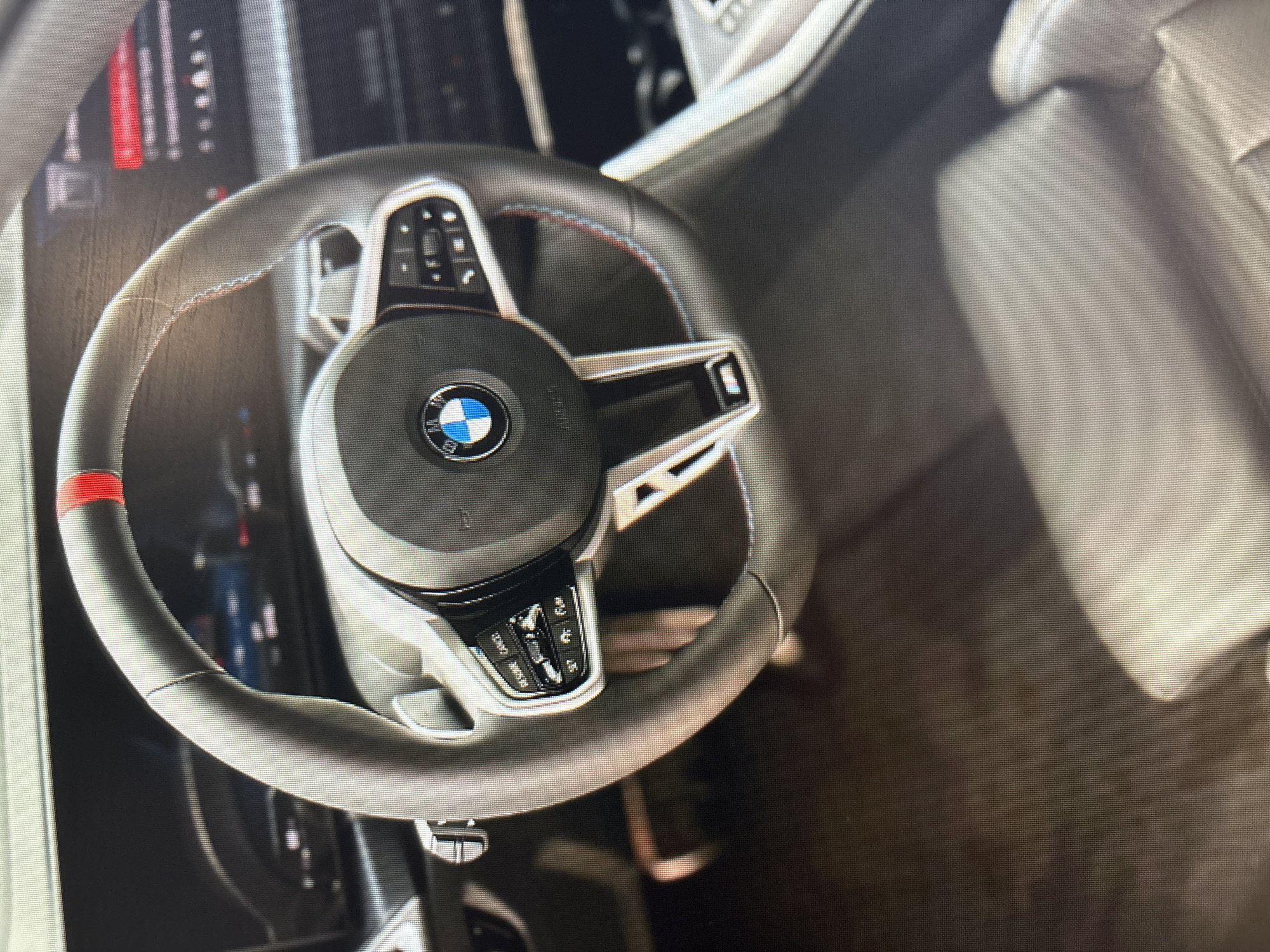 The image shows a close-up of the interior of a BMW M steering wheel, with the dashboard and a portion of a seat visible. The steering wheel is dark gray with red stitching and a red stripe at the 12 o'clock position. The BMW logo is prominently displayed in the center of the steering wheel. Buttons for audio and cruise control are visible on the left side of the steering wheel. The dashboard features a digital display screen on the right, showing red text and indicators. The lighting suggests an indoor setting, possibly a car showroom or a parking garage in Moscow, Russia.