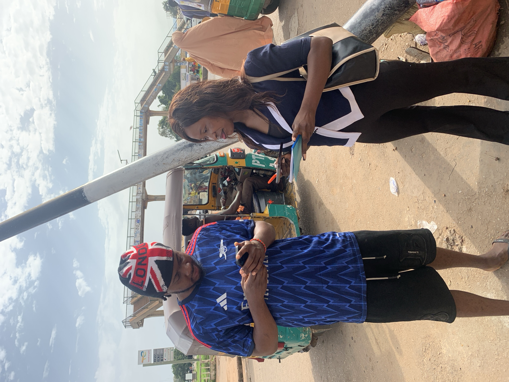 In Gura Top, Nigeria, a man wearing a blue and red Arsenal jersey, black shorts, and a Union Jack beanie, stands looking at his phone. To his right, a woman in a dark blue blazer with white trim, black pants, and a tan and black purse, holds a notepad and pen. They are standing on a dirt road near an auto-rickshaw. In the background, an elevated pedestrian walkway crosses over the road. The sky is partly cloudy.