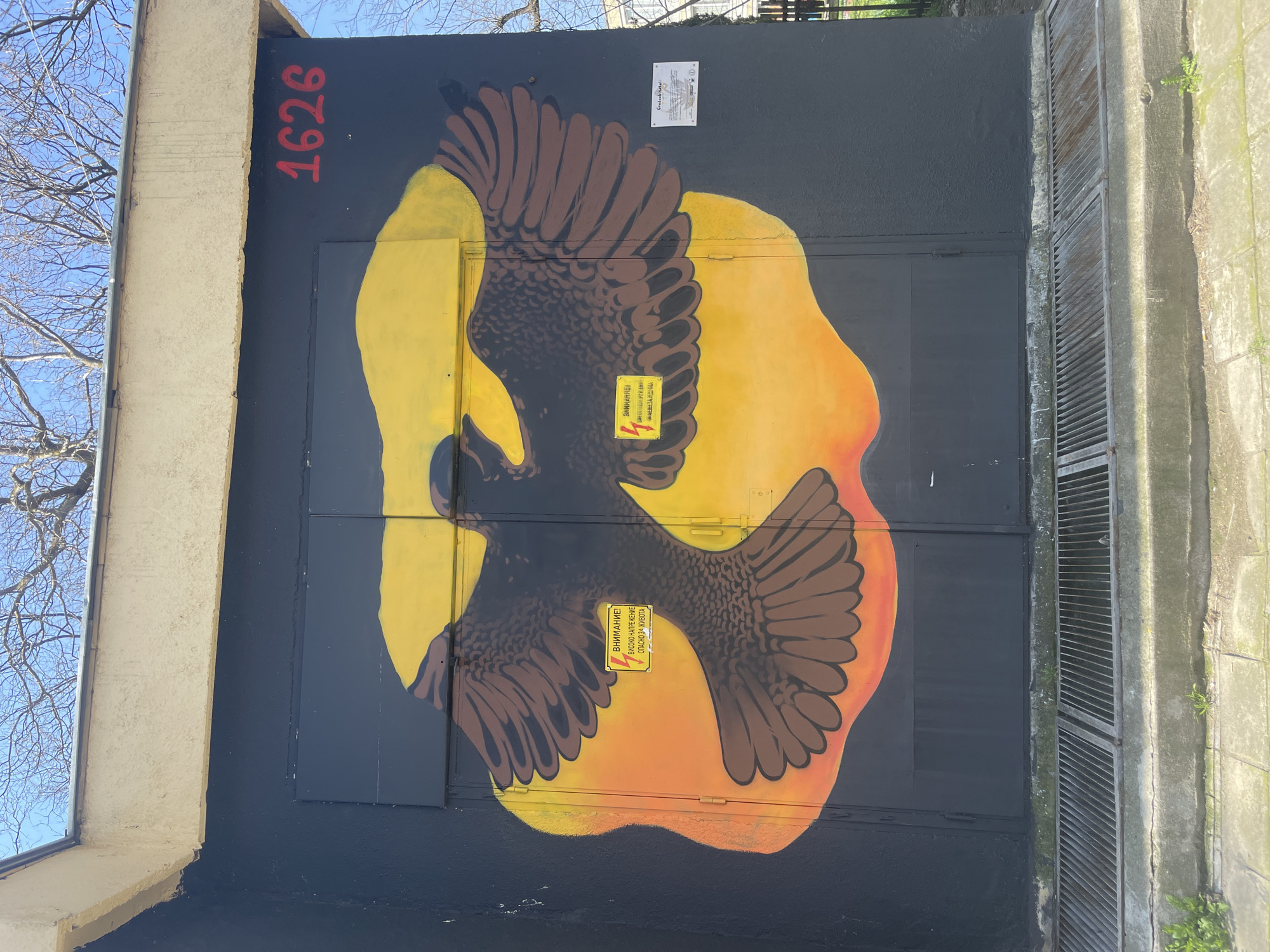 Here's a detailed description of the image:
**Subject:** A mural painted on what appears to be an electrical or utility box. The mural depicts a bird, possibly an eagle or hawk, with outstretched wings silhouetted against a vibrant yellow and orange sun-like shape.
**Location:** Based on the presence of Cyrillic script, the location is likely in Bulgaria. The mural also contains the text “Gradoobitatell” which indicates it could be near the city of Varna.
**Details:**
* **Mural:** The bird is predominantly dark brown with detailed feather patterns. The background sun is painted in bright yellow transitioning to orange towards the edges.
* **Utility Box:** The painting is applied to a metal utility box. Two yellow warning signs with black lettering and a lightning bolt symbol in Cyrillic are affixed to the front of the box. A white certificate-like document is on the upper right.
* **Surroundings:** The box is adjacent to a light-colored stucco building. To the right of the box, there's a concrete structure, likely part of the building's foundation, with ventilation slots.
* **Number:** The number "1626" is painted in red on the left side of the box.
* **Environment:** The sky is visible in the upper-left corner, suggesting an outdoor setting.