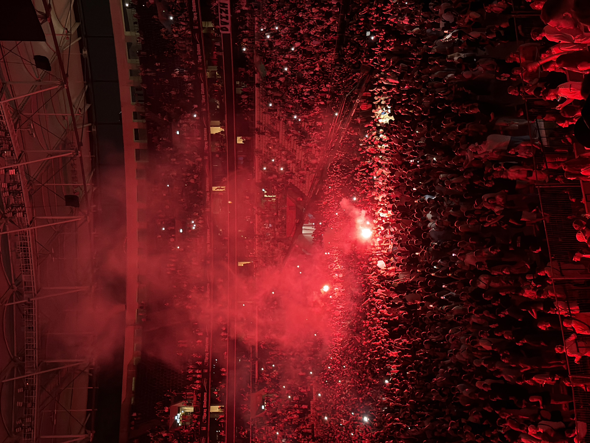 The image displays a large crowd gathered inside Tüpraş Stadyumu, a stadium located in Beşiktaş, Turkey, at night. The scene is illuminated predominantly by a vivid red light and dense red smoke emanating from multiple sources within the audience, suggesting the use of flares or pyrotechnics. Numerous individuals in the packed lower tiers are holding up illuminated smartphones, creating distinct white points of light scattered across the red-lit environment. Banners bearing "Tüpraş Stadyumu" are visible on various levels of the stadium's seating areas. The upper sections of the stadium, including the roof structure and additional seating tiers, are partially visible through the smoke. The crowd appears to be actively engaged, with many individuals facing towards a central point not fully depicted, some with arms raised. Railings and architectural elements of the stadium are discernible through the crowd.