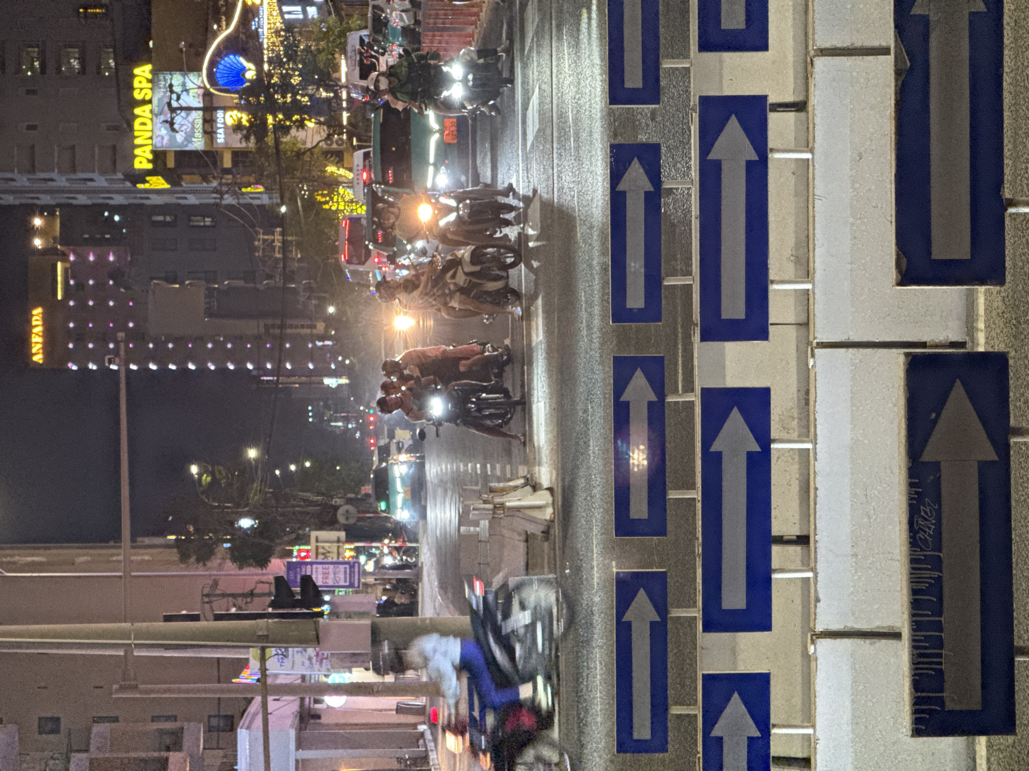 The image captures a bustling night scene on a city street, viewed in a vertical orientation where the road stretches upwards through the frame. On the right, a lane divider features a series of blue rectangular signs with white upward-pointing arrows, clearly indicating the direction of traffic.
The street is filled with a dense flow of vehicles, primarily motorcycles, all moving upwards and away from the viewer. Numerous individuals are visible riding these motorbikes, some alone, others in pairs or small groups, with several wearing helmets. The headlights and taillights of the vehicles create bright points and streaks of light, reflecting on the road surface, which appears either wet or highly reflective.
Along the left side of the road, multi-story buildings are brightly illuminated by various commercial signs. Prominently displayed is a large yellow sign for "PANDA SPA," with a smaller "Massage" sign below it. Further down, another building has a sign that reads "ANFADA." Other distant signs include "SEA FOOD" and "FREE W." A distinctive blue, shell-shaped illuminated sculpture is also visible near the "Panda Spa" sign. In the lower left, a person on a bicycle can be seen, also moving in the direction of traffic. The overall scene conveys the energetic and busy atmosphere of an urban center at night, typical of Đà Nẵng, Vietnam.