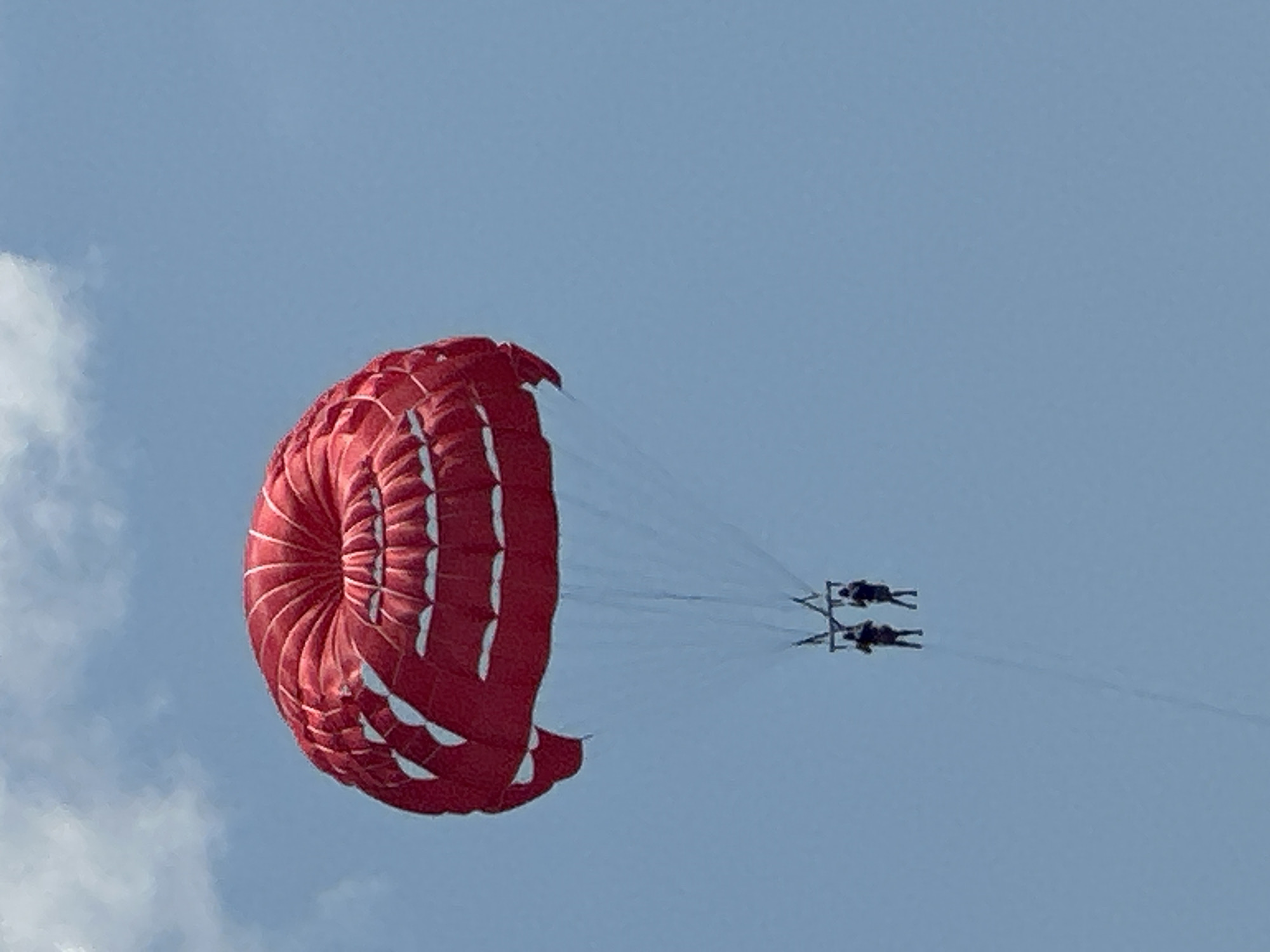 This image shows two people parasailing in the sky above Boreti, Montenegro. The parasail is red and white, and the people are suspended from it by ropes. The sky is blue with some white clouds.