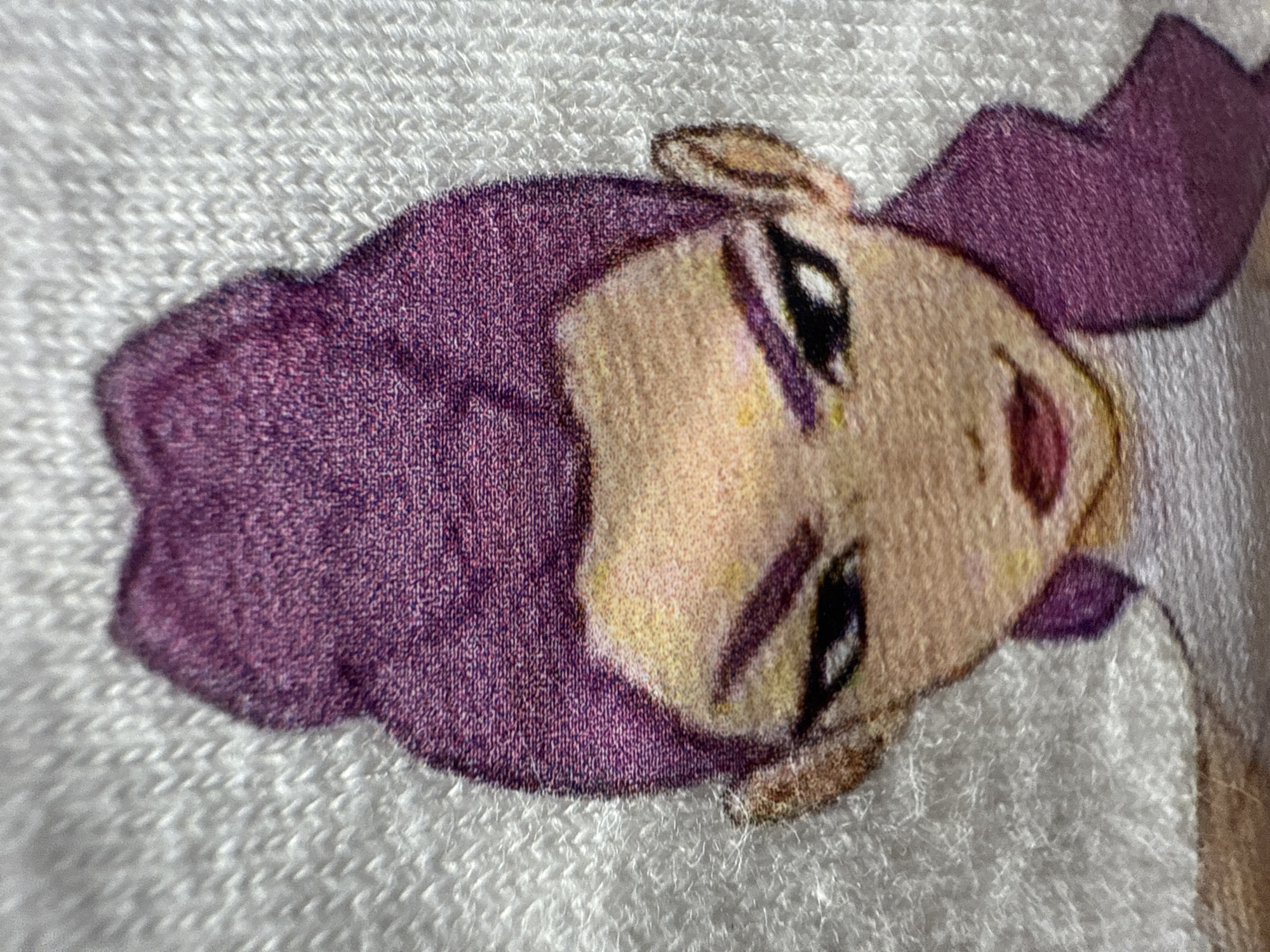 A close-up, rotated image captures a vibrant cartoon character's face printed on a white, textured fabric, likely a towel or similar soft material. The character, seen in profile, features striking purple hair that frames a light-skinned face with yellow and pink undertones. Large, expressive eyes with black pupils and subtle purple detailing suggest a lively or mischievous gaze, complemented by a small, reddish mouth indicating a slight smile. The print is integrated into the fabric, revealing the underlying looped texture of the white material, characteristic of terrycloth. The image is oriented with the character's head tilted to the side, looking upwards to the right. No specific activity or event is taking place beyond the presentation of this printed graphic. There is no visible text. The notable details are the clear fabric texture and the bright, cartoonish aesthetic of the character.
