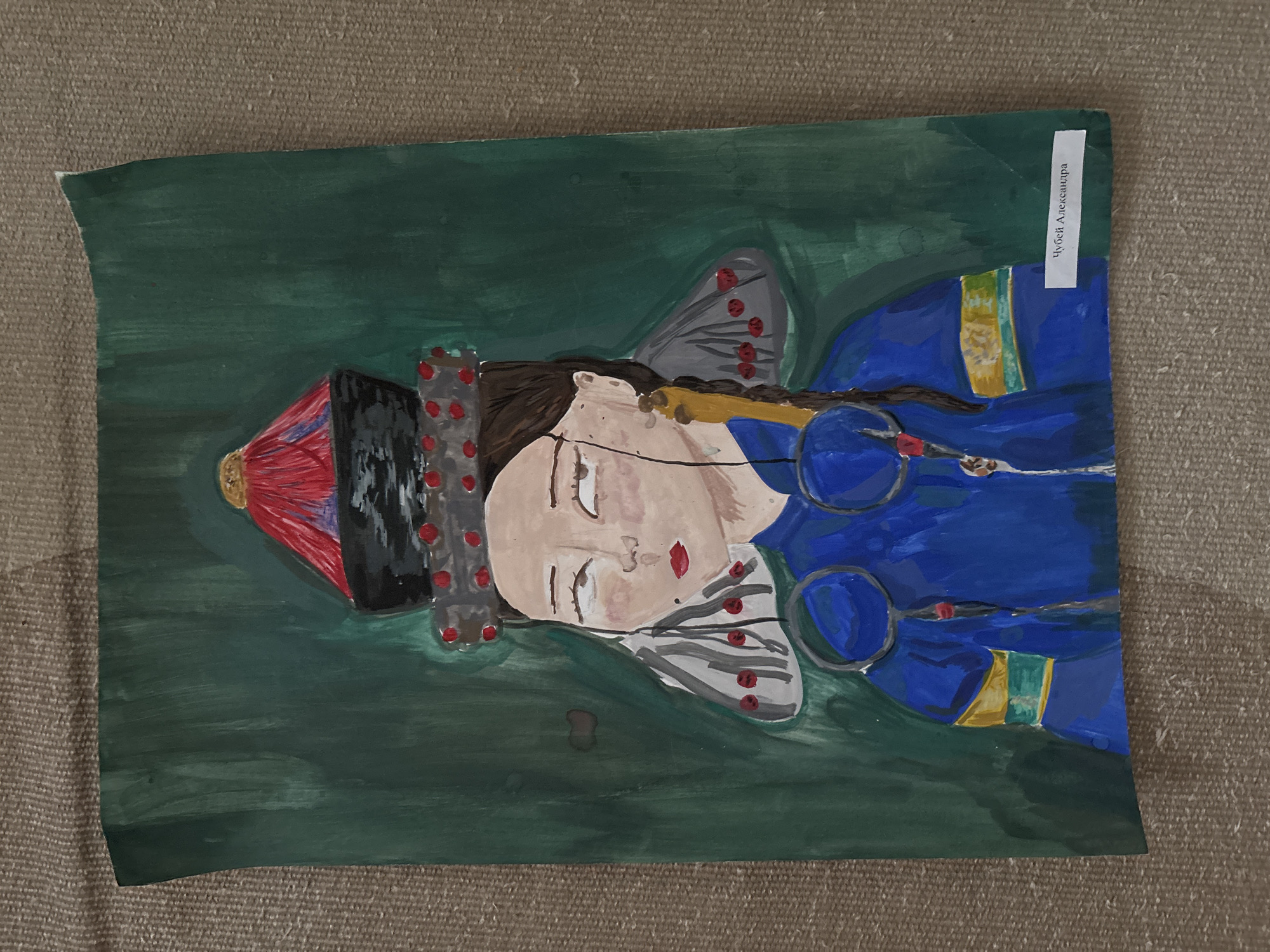 The image shows a child's painting of a woman in traditional Russian clothing. The woman has fair skin, closed eyes, and red lips. She wears a dark headdress with red decorations, a blue dress with yellow and green accents, and fan-shaped ornaments by her face. The background is a dark green color. A white label with Cyrillic text ("Чубей Александра" - Chubey Alexandra) is attached to the upper right corner of the painting. The painting is laying on a beige surface. The subject is dressed in traditional Russian clothing so the painting is likely related to that cultural theme. Saint Petersburg is not visible in the painting itself.