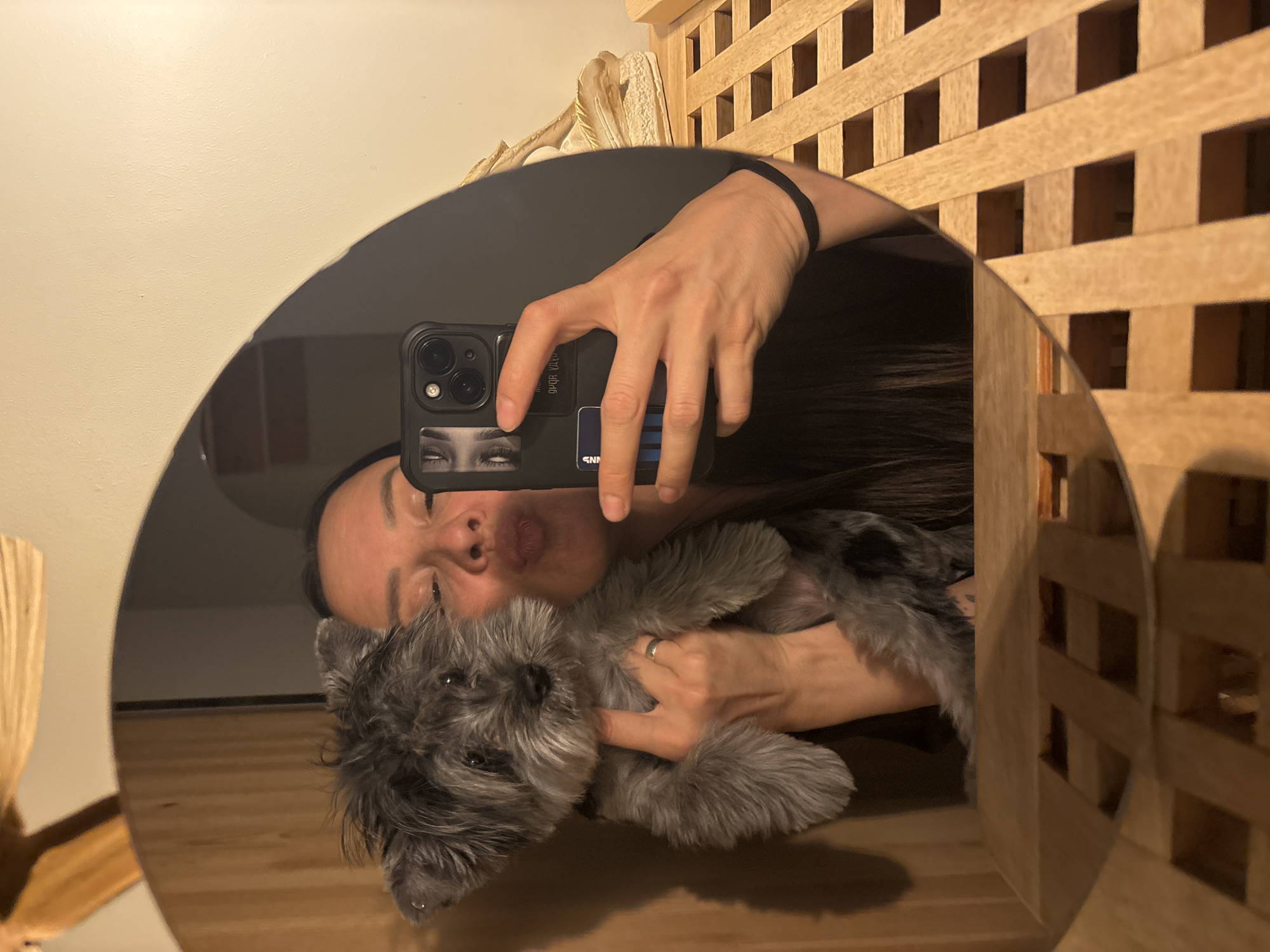 A selfie, taken in a round mirror, shows a person holding a gray dog, possibly a terrier mix. The person holds a black smartphone with a sticker of a pair of eyes and a blue rectangular sticker on the back. The person has long, dark hair and is wearing a black headband. The dog has shaggy fur and is looking at the camera. The environment includes a light wood-toned wall with a lattice-like design, and a light-colored wall and ceiling are visible in the mirror as well. No evidence to indicate the location is Chaikovsky, Russia.