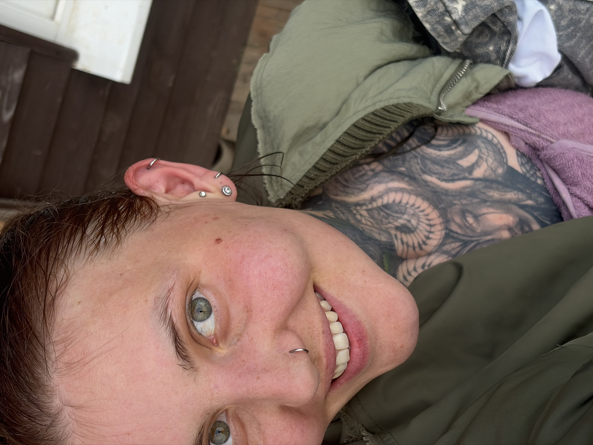 This is a close-up, vertically oriented photograph of a person lying down. The subject's face is tilted upwards and to the left, with their eyes open and looking towards the camera. They have light-colored eyes, and their mouth is open in a smile, revealing teeth. The person has several piercings: a hoop in their nose, and multiple small rings and studs in their ear. Their hair is dark brown and appears slightly damp.
The person is wearing an olive-green jacket, and part of a purple, fuzzy blanket is visible. A large, intricate tattoo is visible on their chest and shoulder, featuring what appears to be a serpent or dragon. The background is blurred, but suggests an outdoor setting with wooden planks and a white structure. The lighting suggests it is daytime, likely overcast. The overall impression is one of casual relaxation. There is no visible text in the image. The provided location context is Malyi Bukor, Russia.