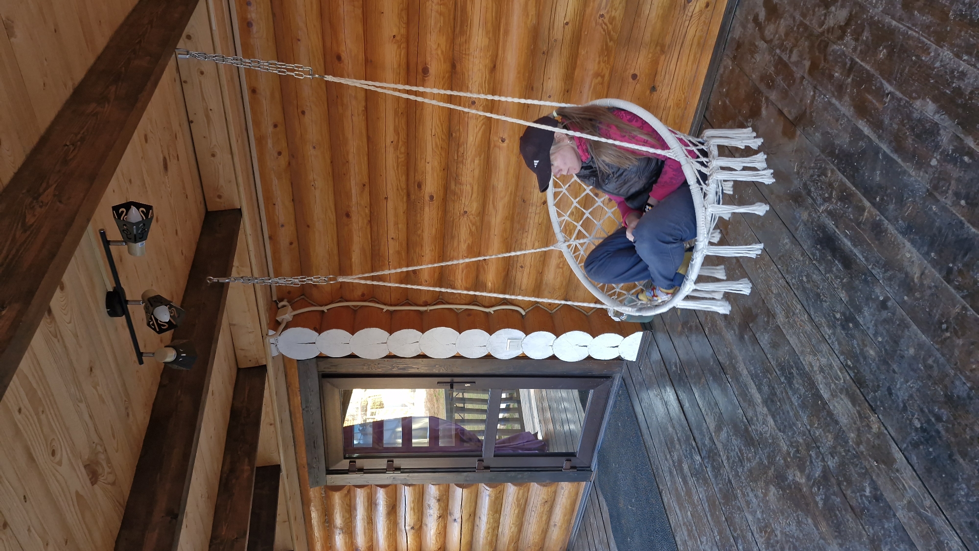A woman sits in a hanging chair indoors, the interior is wood-paneled. The woman is wearing a black baseball cap, a pink shirt, a dark vest, and blue jeans. She appears to be looking down. The chair is made of white rope and has fringe hanging from the bottom. The chair hangs from the ceiling by ropes and chains. Above the woman is a wooden ceiling, and next to her is a window. Below the chair is a wooden floor. A light fixture is also visible on the ceiling. It is impossible to verify the location as Otchor, Russia.