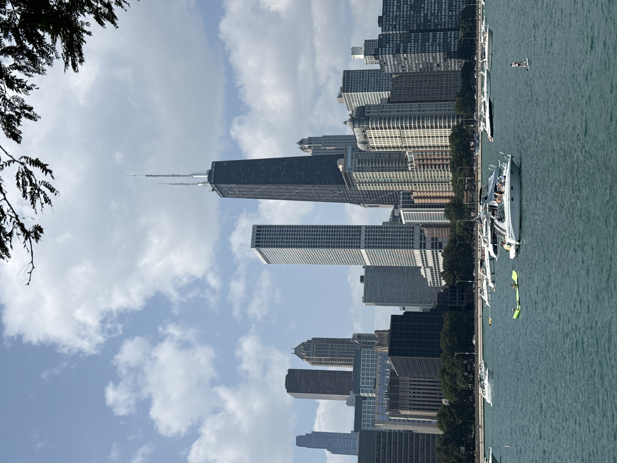The image depicts a view of the Chicago skyline from Lake Michigan on a clear day with scattered clouds. In the foreground, the turquoise water of the lake is dotted with several white recreational boats of various sizes. One boat in the center has several people on board. To the left, a green inflatable float is being used by a person lying on their stomach. Further in the distance, a person is seen paddleboarding.
The middle ground features a line of trees separating the lake from the city. The background is dominated by a dense collection of skyscrapers. The most prominent building is the distinctive black John Hancock Center, which stands tall among other modern and older architectural styles. Other notable buildings include various glass and concrete structures. The sky above is a bright blue, with fluffy white clouds providing a contrast to the urban landscape. The overall scene captures a typical summer day in Chicago, with people enjoying the lake and the city's iconic architecture visible.