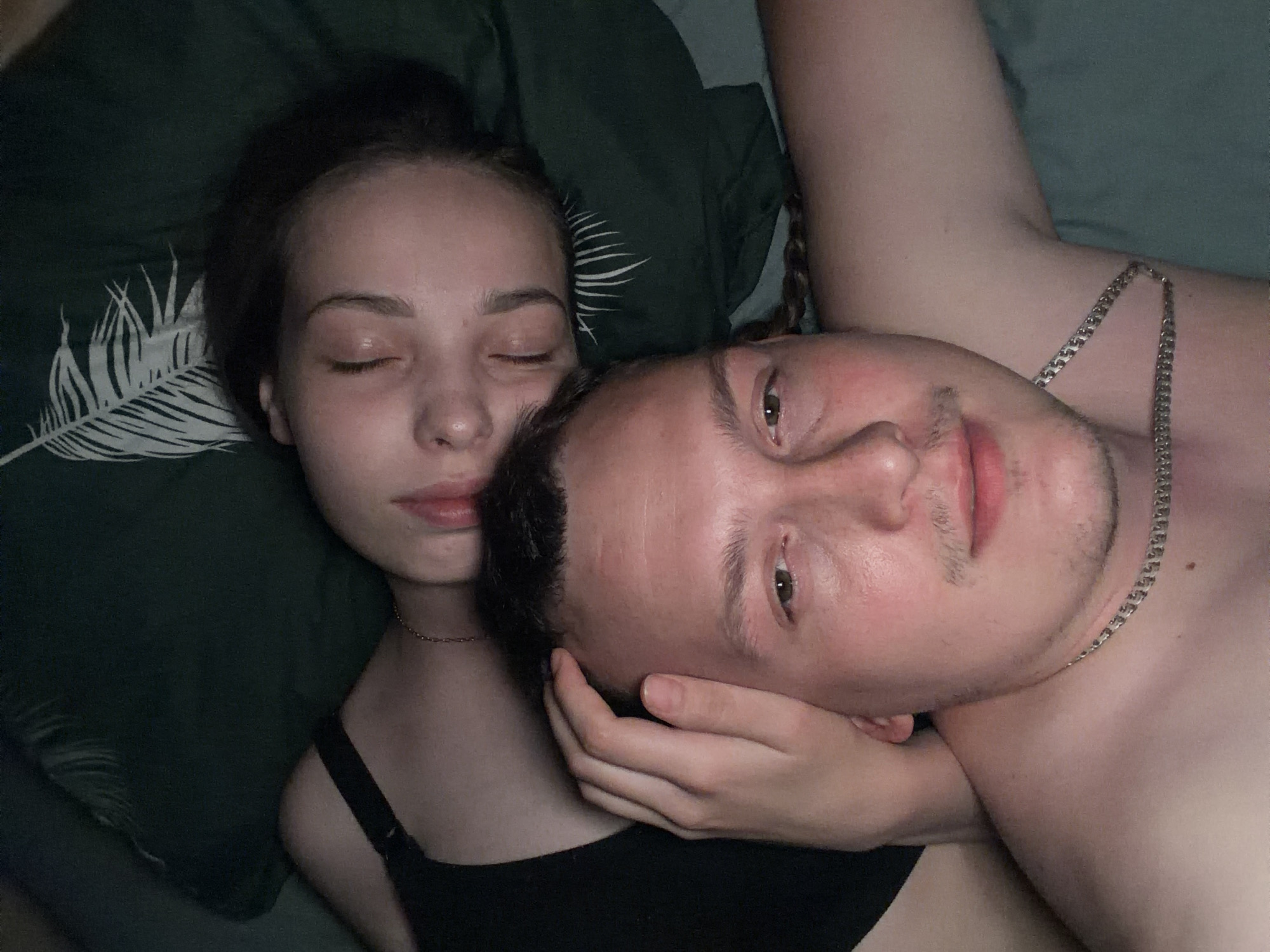 A close-up, overhead shot shows a couple lying down, possibly in bed, with their faces close together. The person in the foreground, appearing to be male, is shirtless and looking towards the camera, with a silver chain around their neck. The person in the background, appearing to be female, is lying on their side, with their eyes closed, and their head resting on a dark green pillow with a white feather pattern. Their left hand is touching the man's forehead. The overall lighting is dim, suggesting an indoor setting. No specific location in Chaikovsky, Russia can be identified from the image.