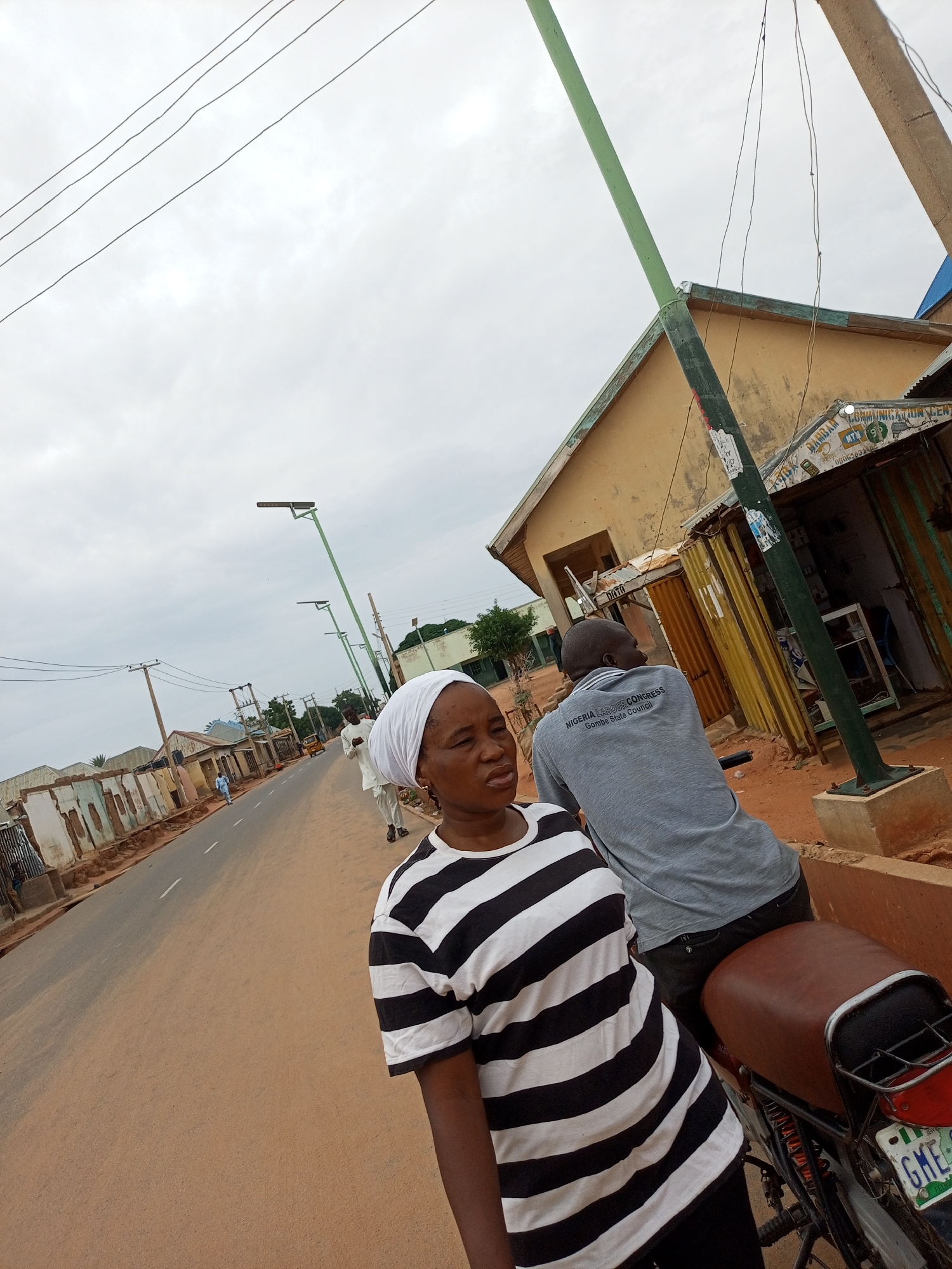 In Gombe, Nigeria, a woman in a black and white striped shirt and white headscarf stands in the foreground, looking to her left. Behind her, a man wearing a grey t-shirt with "NIGERIA LABOUR CONGRESS Gombe State Council" printed on the back, sits on a brown motorcycle. The motorcycle has a license plate that reads "GME 466 AB". They are positioned on a dusty roadside next to a building with a corrugated metal roof and yellow walls. The street is paved with asphalt and lined with utility poles and streetlights. In the background, more buildings and a few people are visible, suggesting a bustling urban environment. The sky is overcast, indicating a cloudy day.